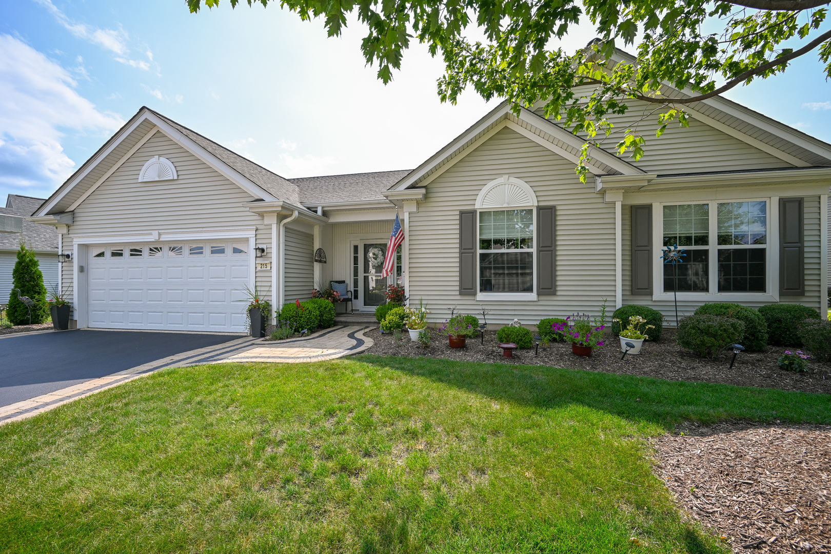 Beige single-story home with a two-car garage, landscaped flower beds, curved walkway, and American flag by the entrance.