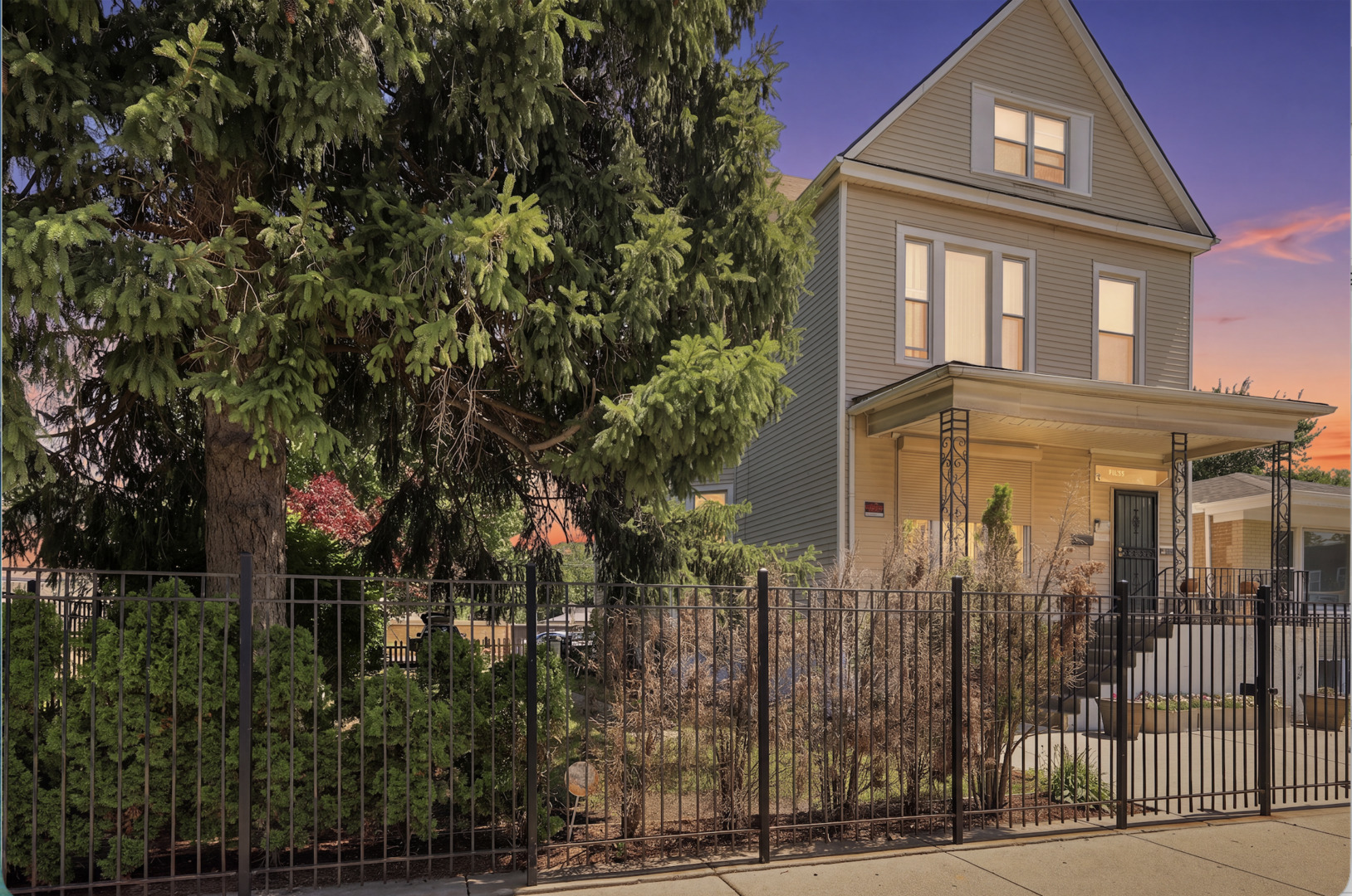 Two-story beige house with a steep gable and covered porch behind a black metal fence at dusk.