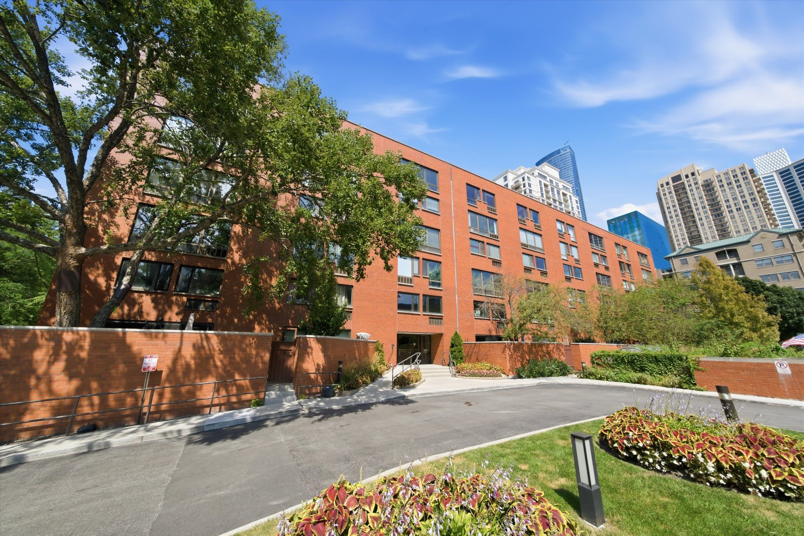Red-brick residential building with mature trees, landscaped grounds, and downtown high-rises behind it.