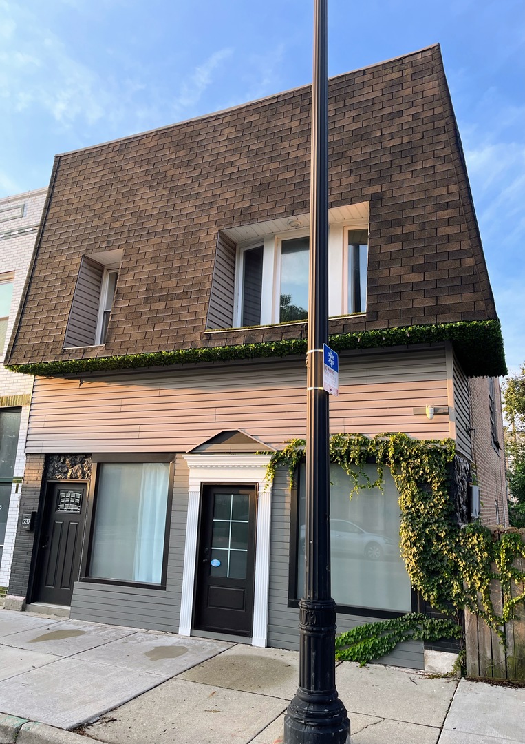 Two-story streetfront building with a dark shingled upper facade, storefront windows, and ivy along one side.