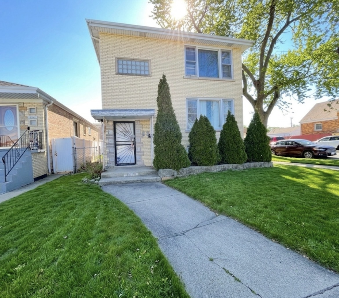 Two-story brick home with a security door, trimmed evergreens, and a concrete walkway through the front lawn.
