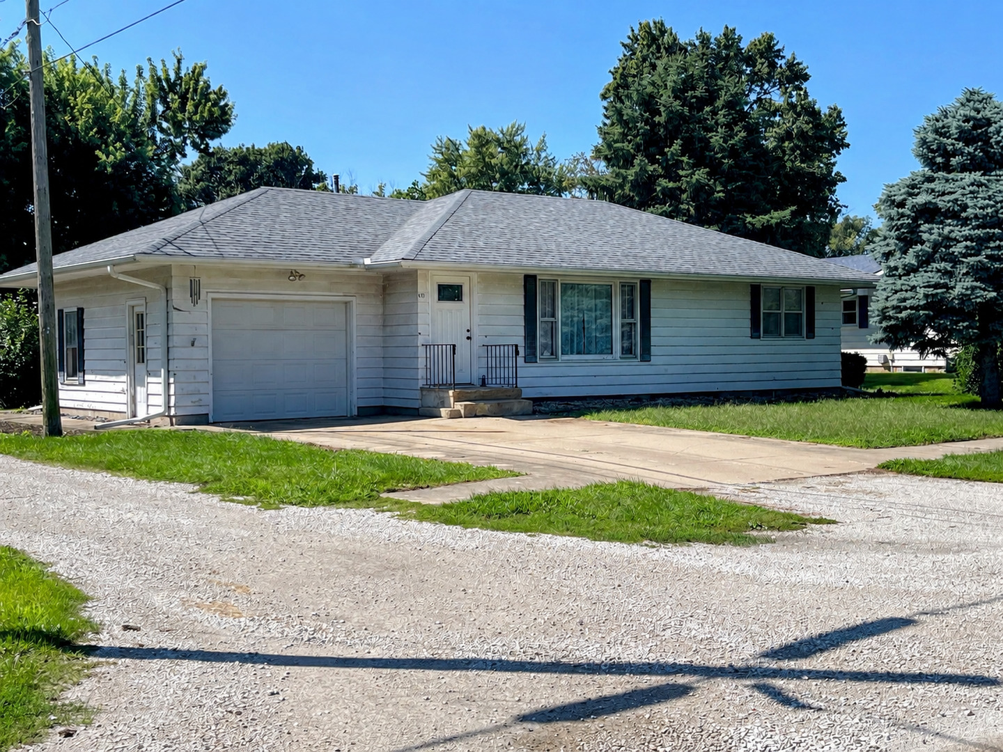 Single-story white house with an attached garage, broad gravel driveway, and mature trees.