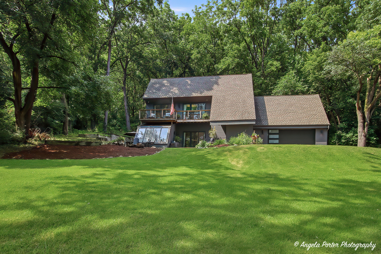 Modern multi-level house with an upper balcony and attached garage, surrounded by trees and a broad lawn.