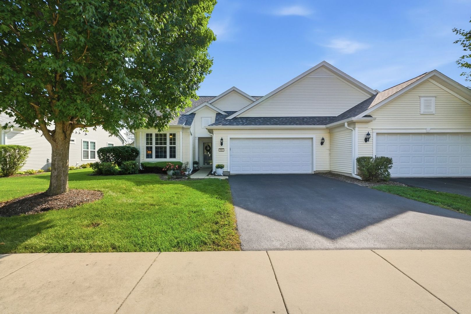 Single-story light-colored home with a two-car garage, broad driveway, landscaped lawn, and large shade tree.