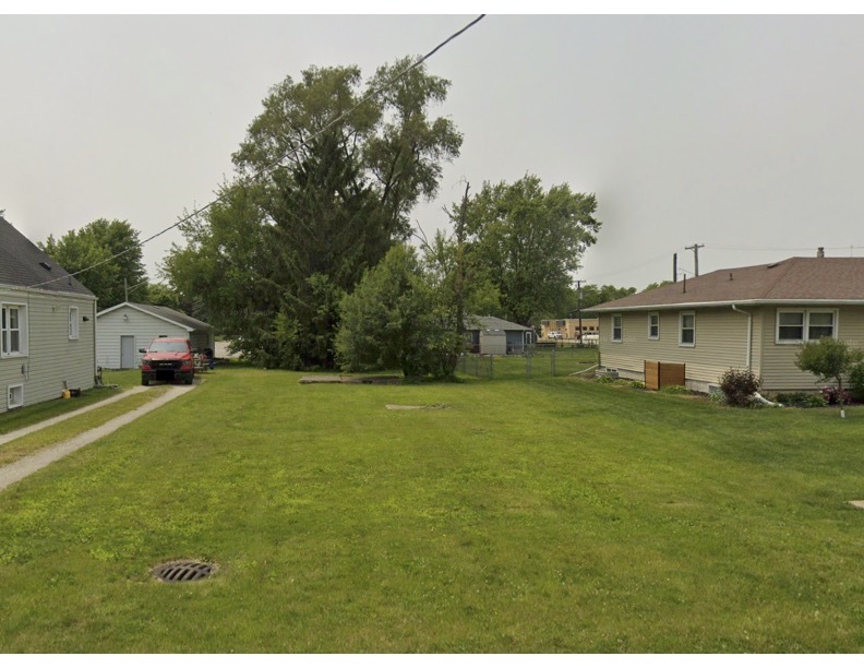 Grassy neighborhood yard between single-story homes, with mature trees and a red vehicle.