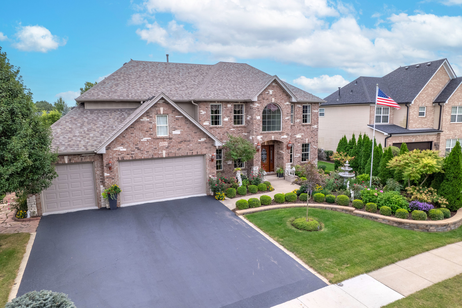 Two-story brick home with a three-car garage, landscaped garden, and American flag.