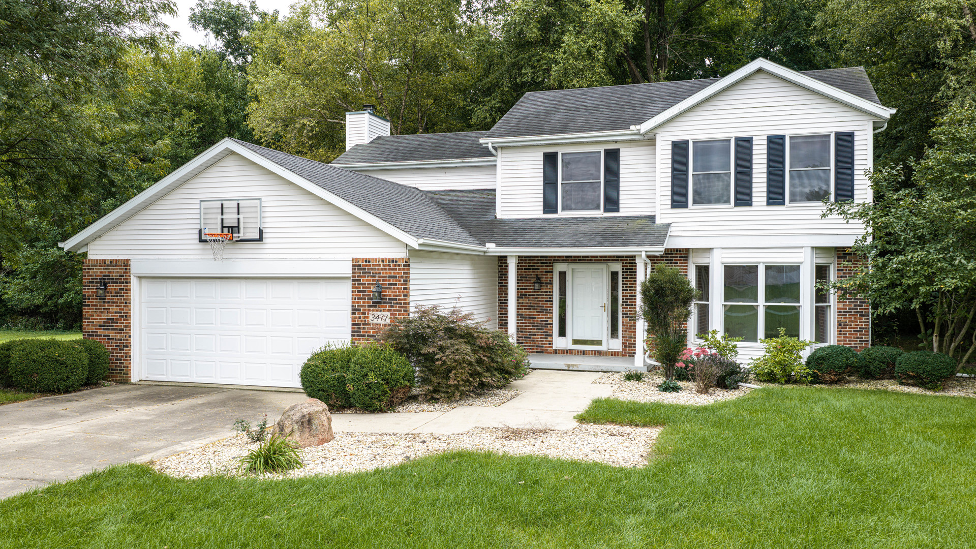 Two-story white-sided home with brick accents, a basketball hoop, and a landscaped front lawn.