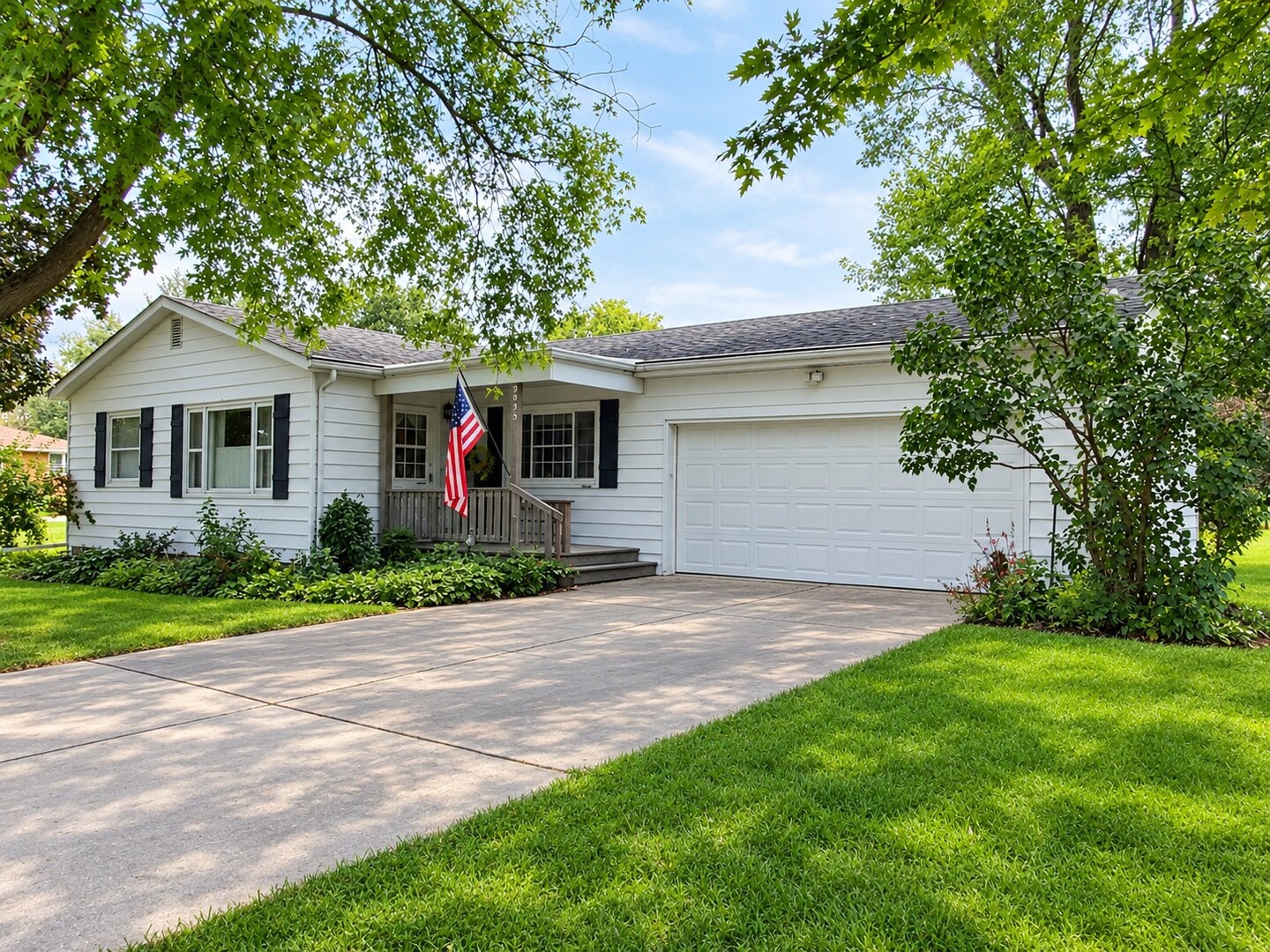 White single-story home with black shutters, an attached garage, and an American flag by the porch.