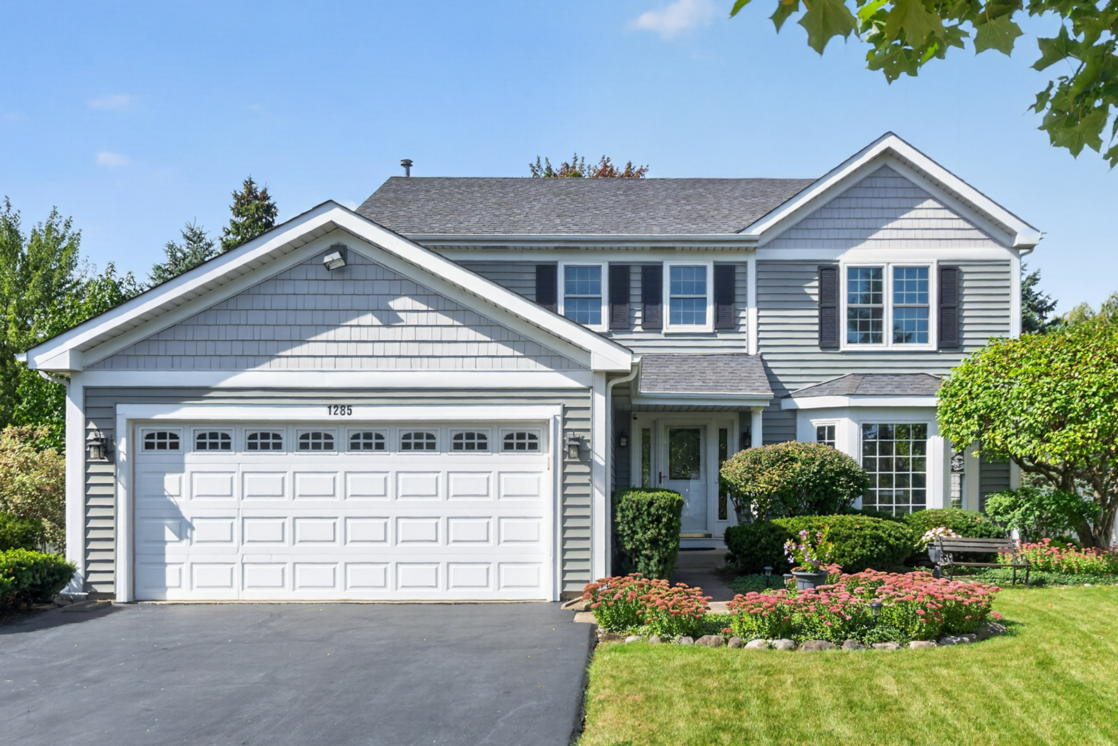 Two-story gray house with a double garage, white trim, dark shutters, and flower beds.