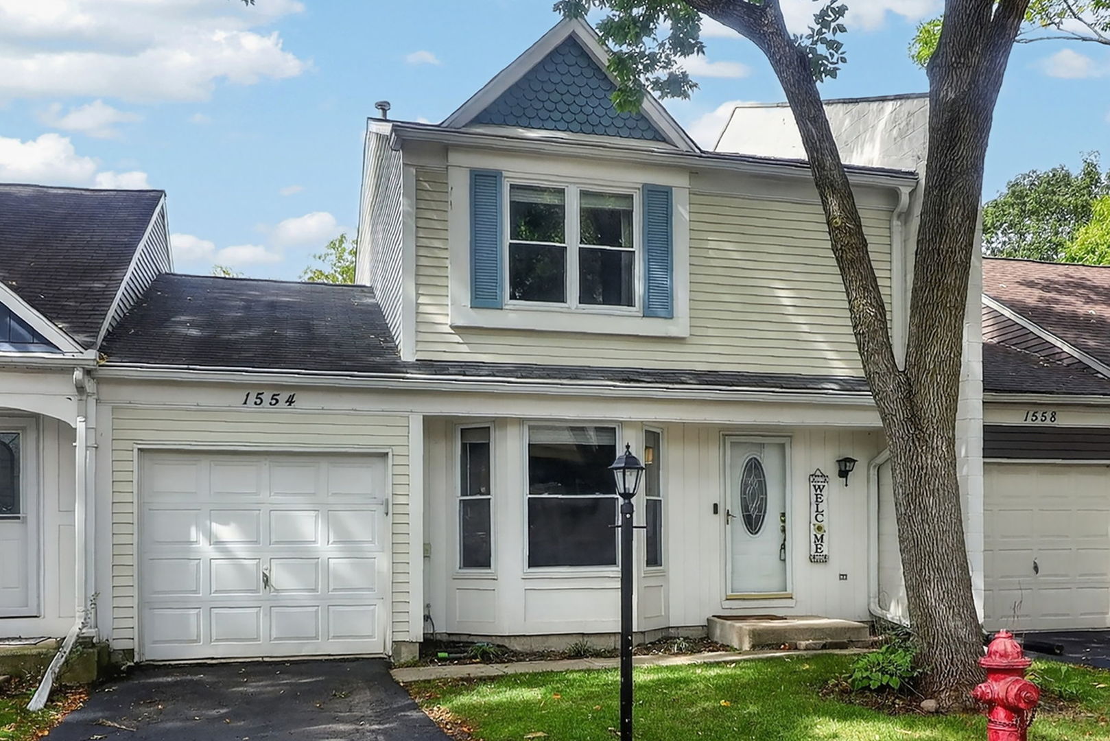 Cream two-story attached home with blue shutters, a front-facing garage, and mature trees.