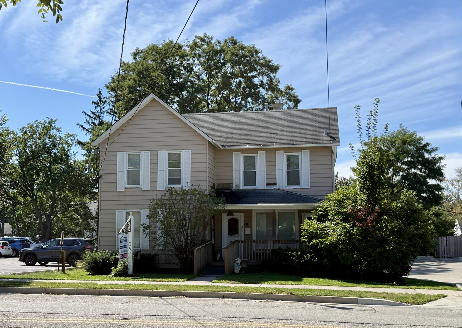 Beige two-story house with white shutters, a covered porch, and mature shrubs along the front.