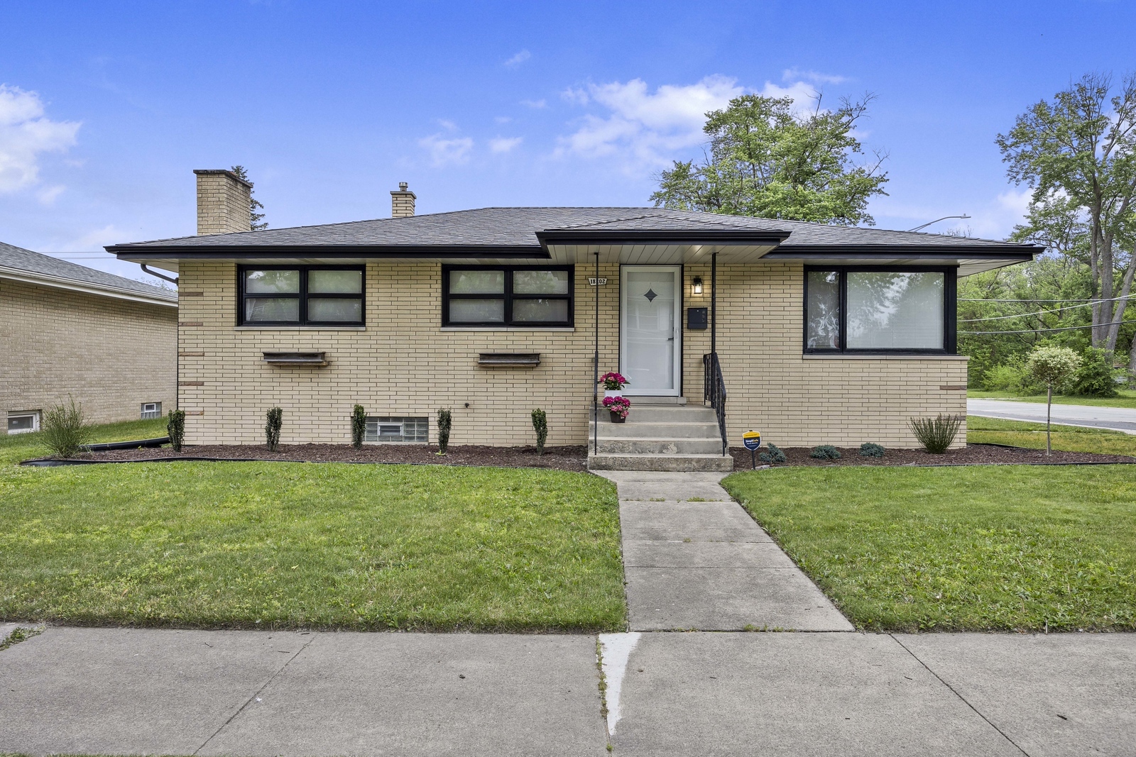 Single-story beige brick home with a broad lawn, dark roof, and concrete walkway to the front steps.