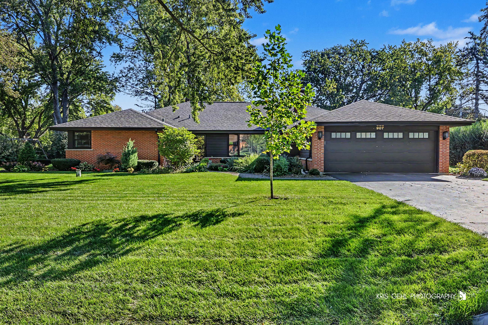 Single-story brick home with a dark two-car garage, landscaped yard, and broad green lawn.