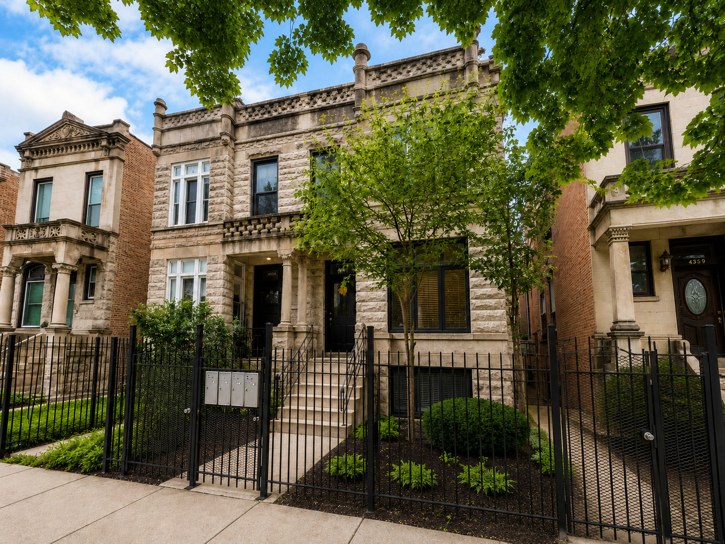 Historic stone home with carved facade, front steps, and black iron fence beneath leafy trees.