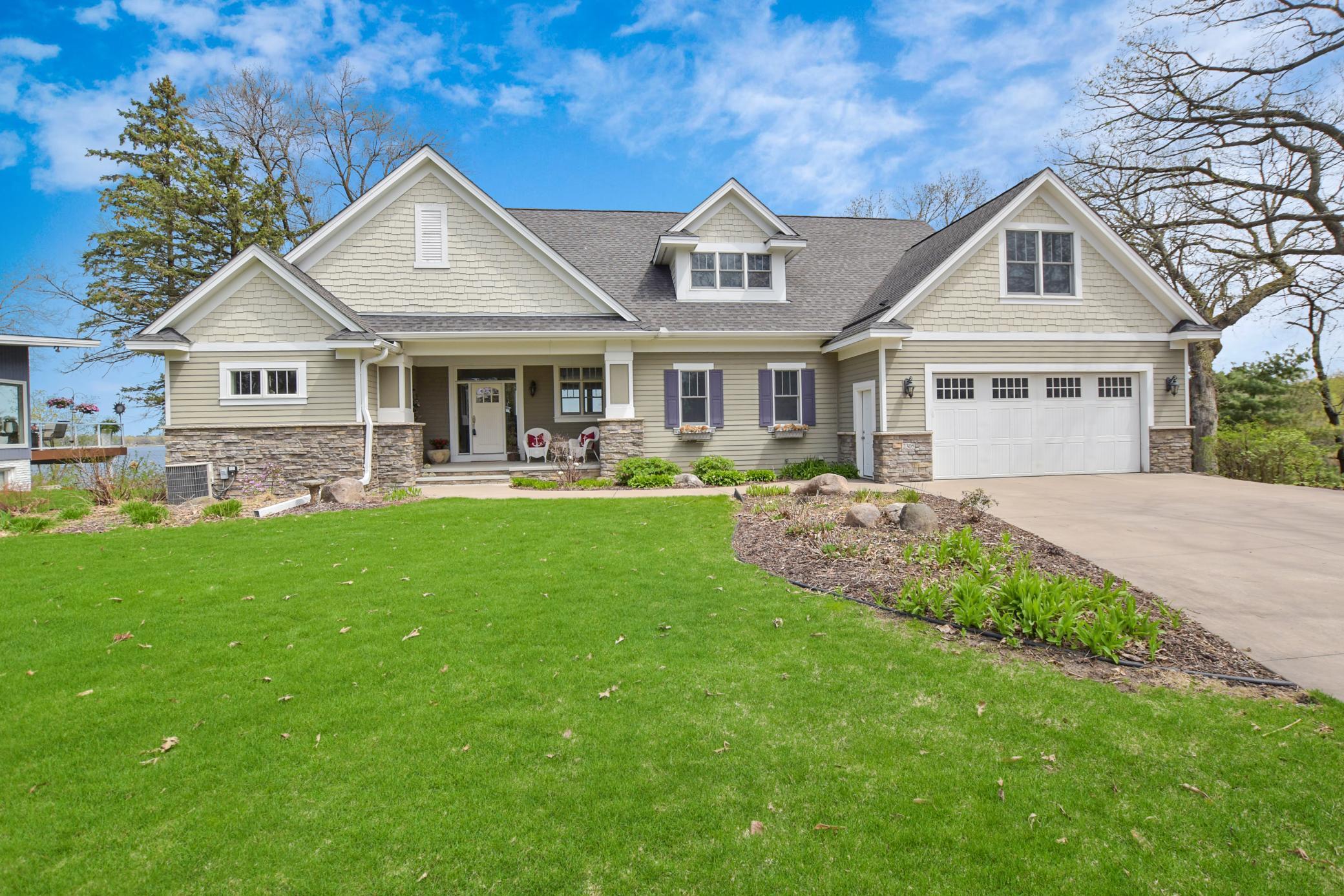 Light-gray gabled home with stone accents, a covered porch, two-car garage, and a glimpse of water nearby.
