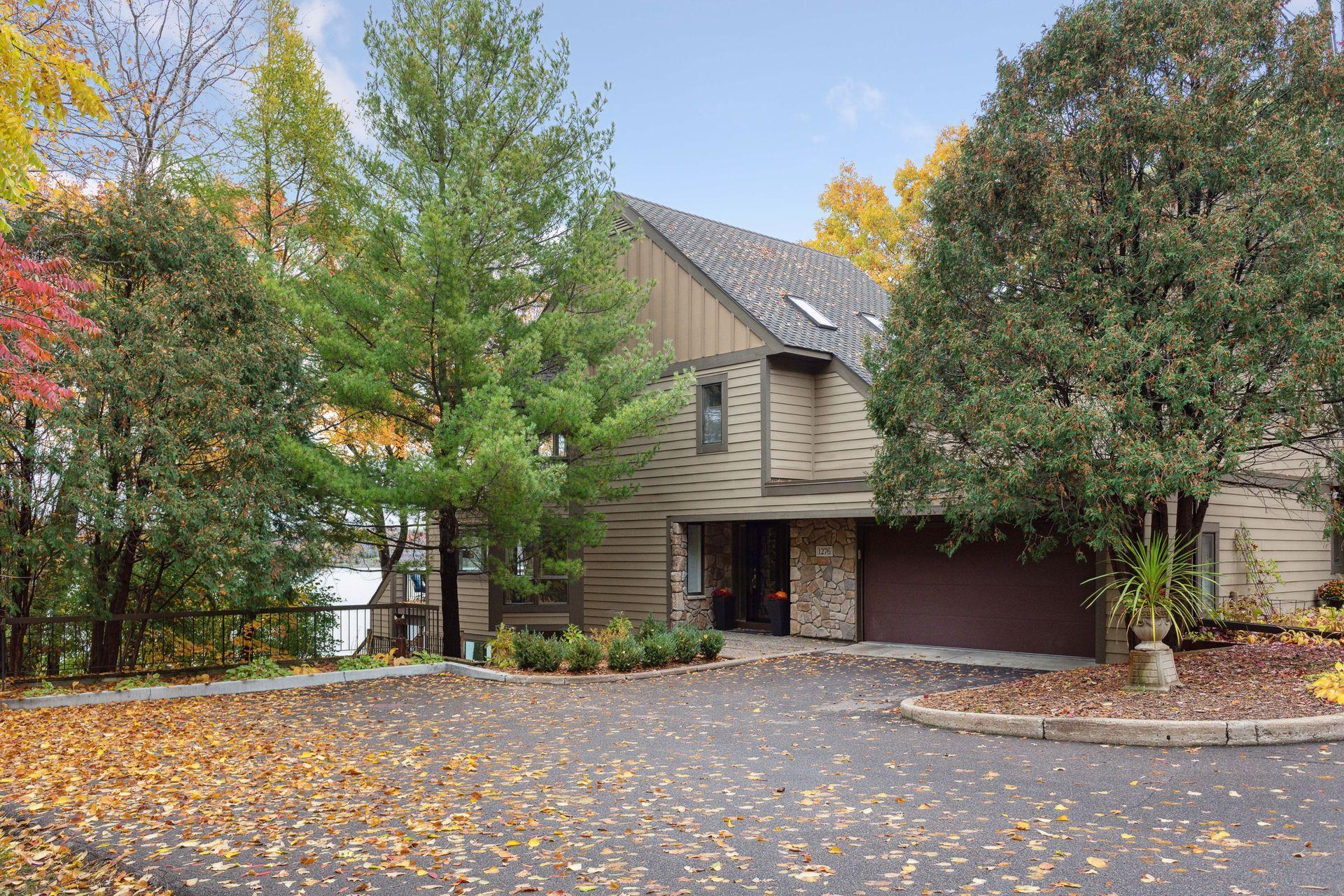 Tan-sided house with a stone entry, partly hidden by trees beside the water in autumn.