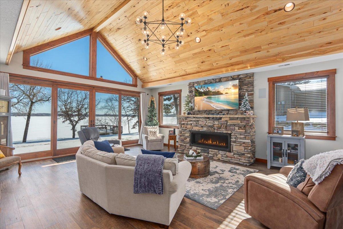 Living room with a vaulted wood ceiling, stone fireplace, and large windows overlooking a snowy lake.