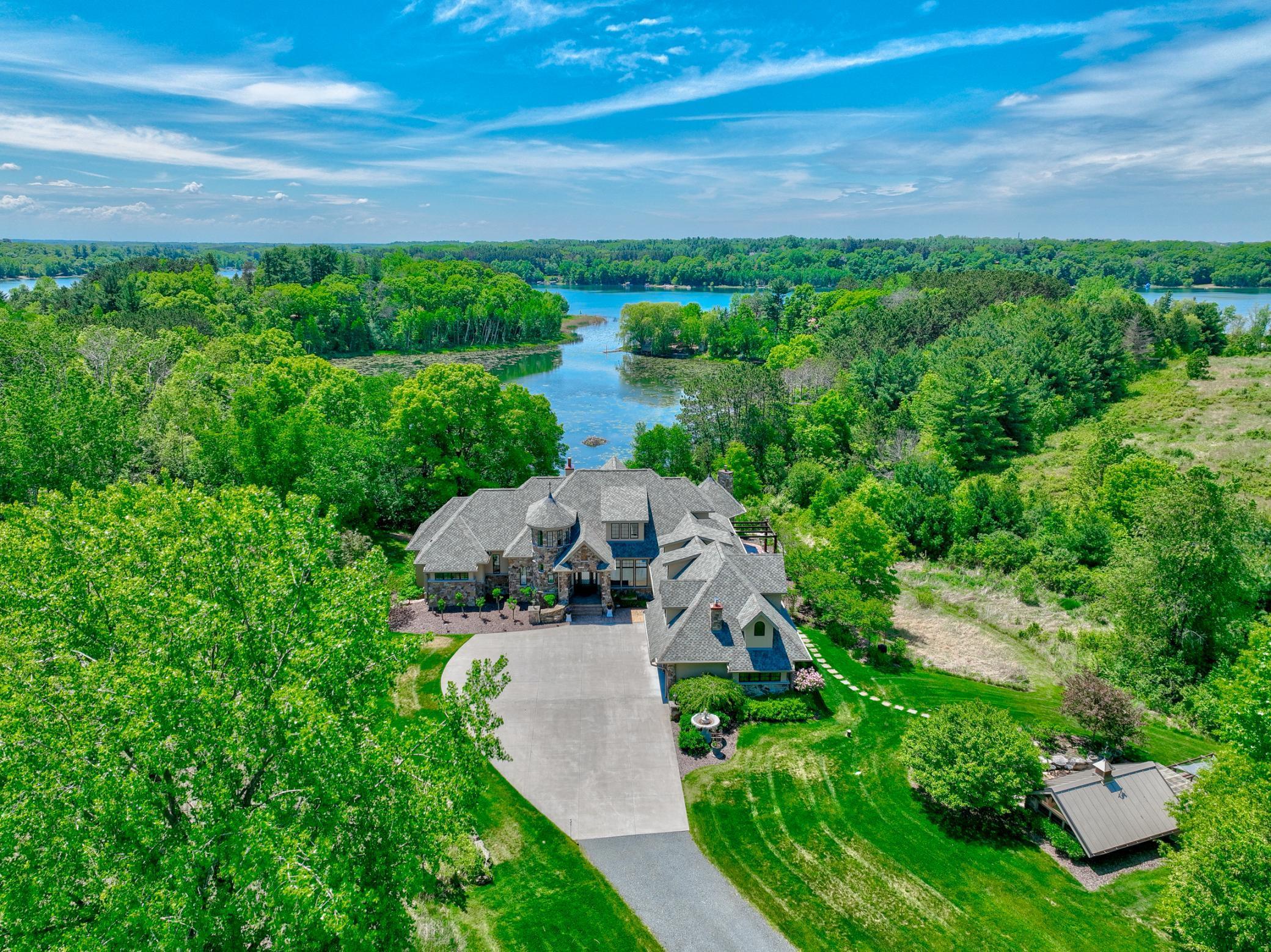 Aerial view of a large stone house surrounded by trees beside a lake, with a broad driveway and landscaped lawn.