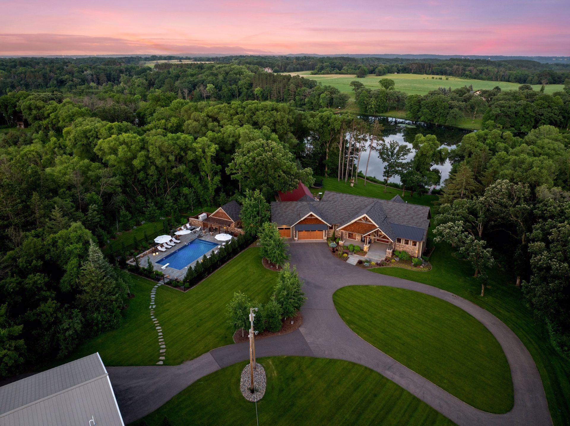 Aerial view of a wooded home with a pool and pond at dusk.