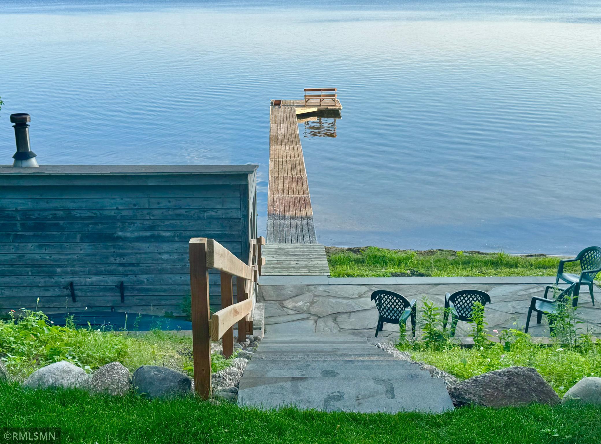 Lakeside dock extending into calm water beside a stone patio and outdoor chairs.