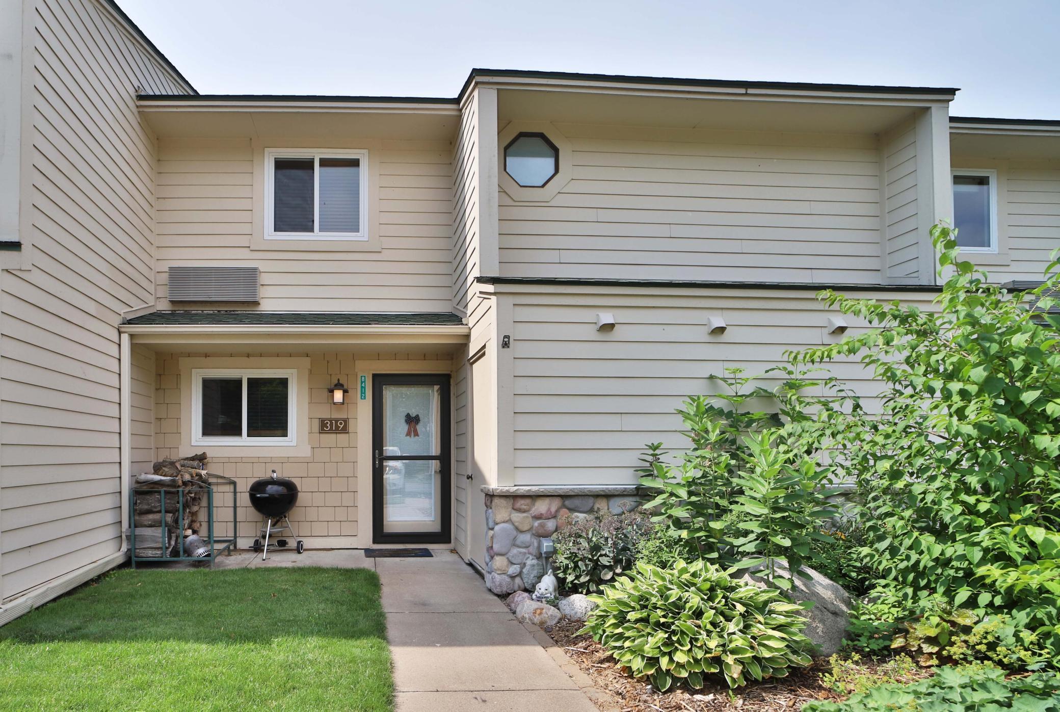 Beige two-story home with a covered entrance, front walkway, lawn, and landscaped shrubs.