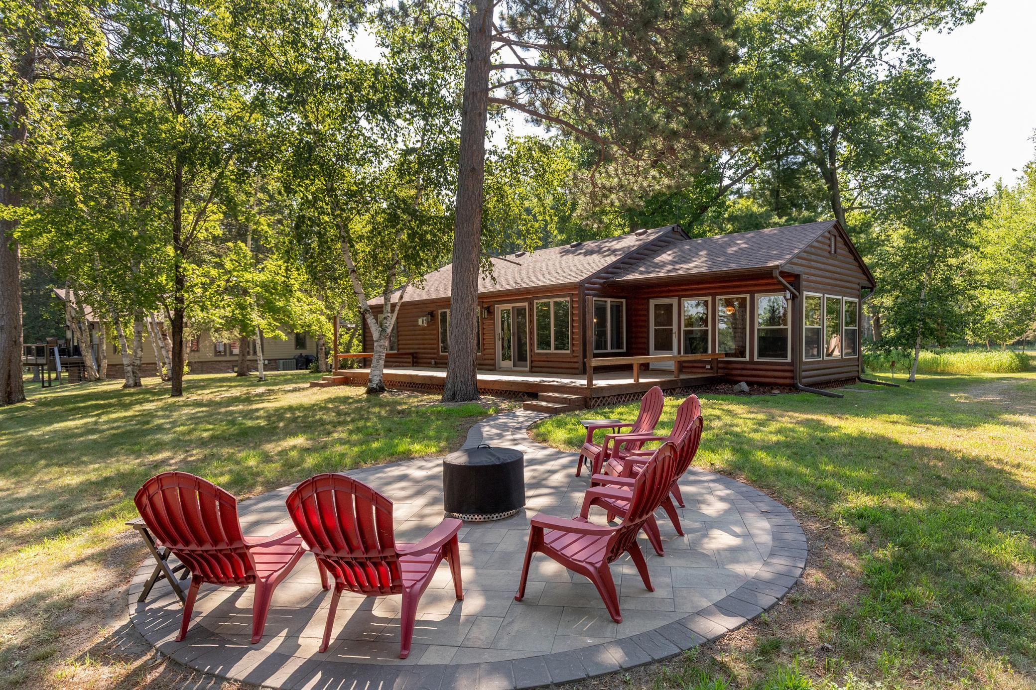 Log-sided cabin behind a circular patio with red Adirondack chairs and a fire pit, shaded by mature trees.