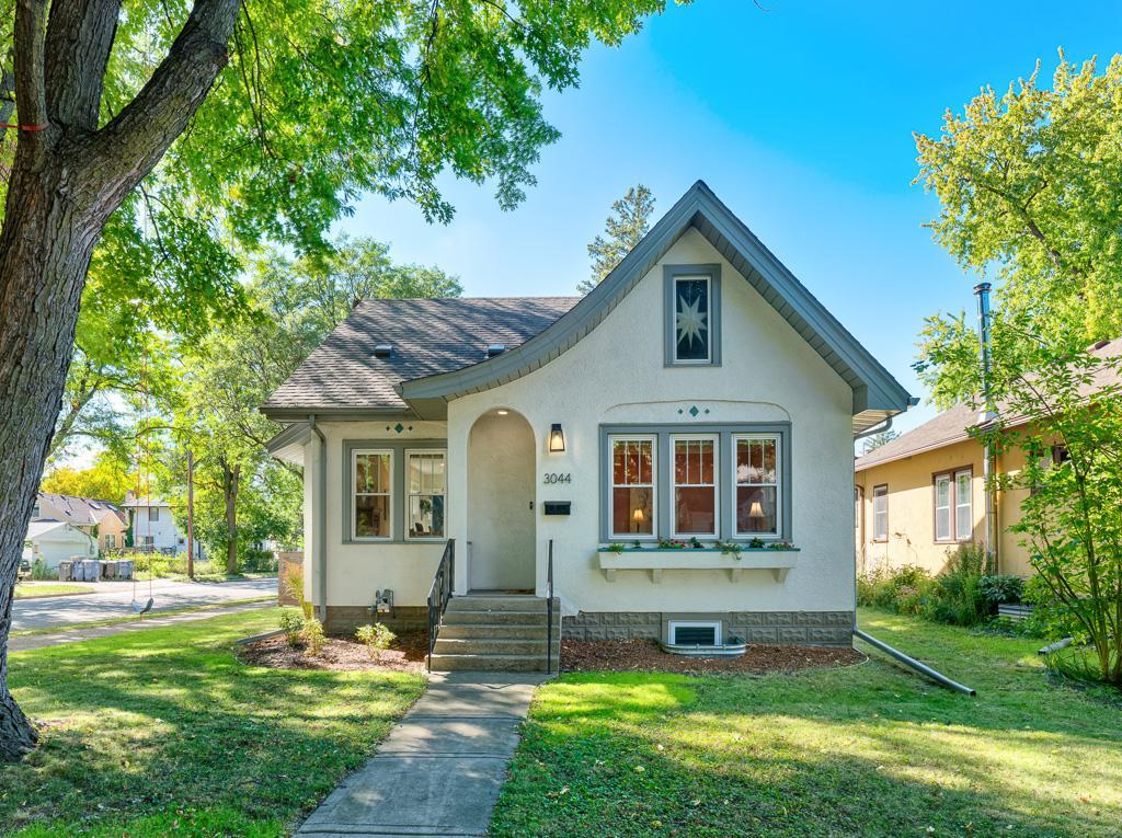 White stucco bungalow with a steep gable, arched entry, flower box, and leafy tree framing the yard.