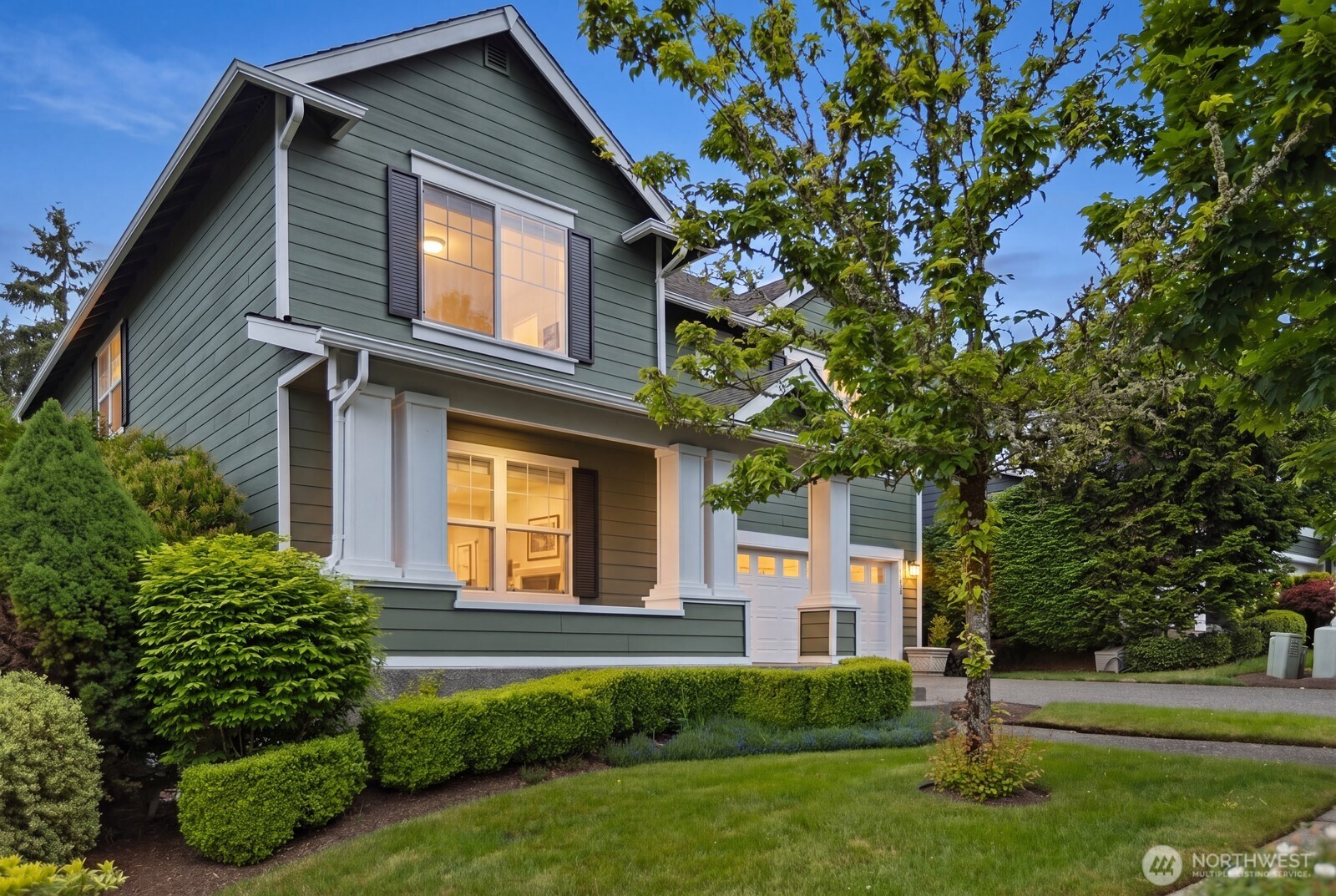 Two-story green home with white trim, a front porch, attached garage, and landscaped yard.