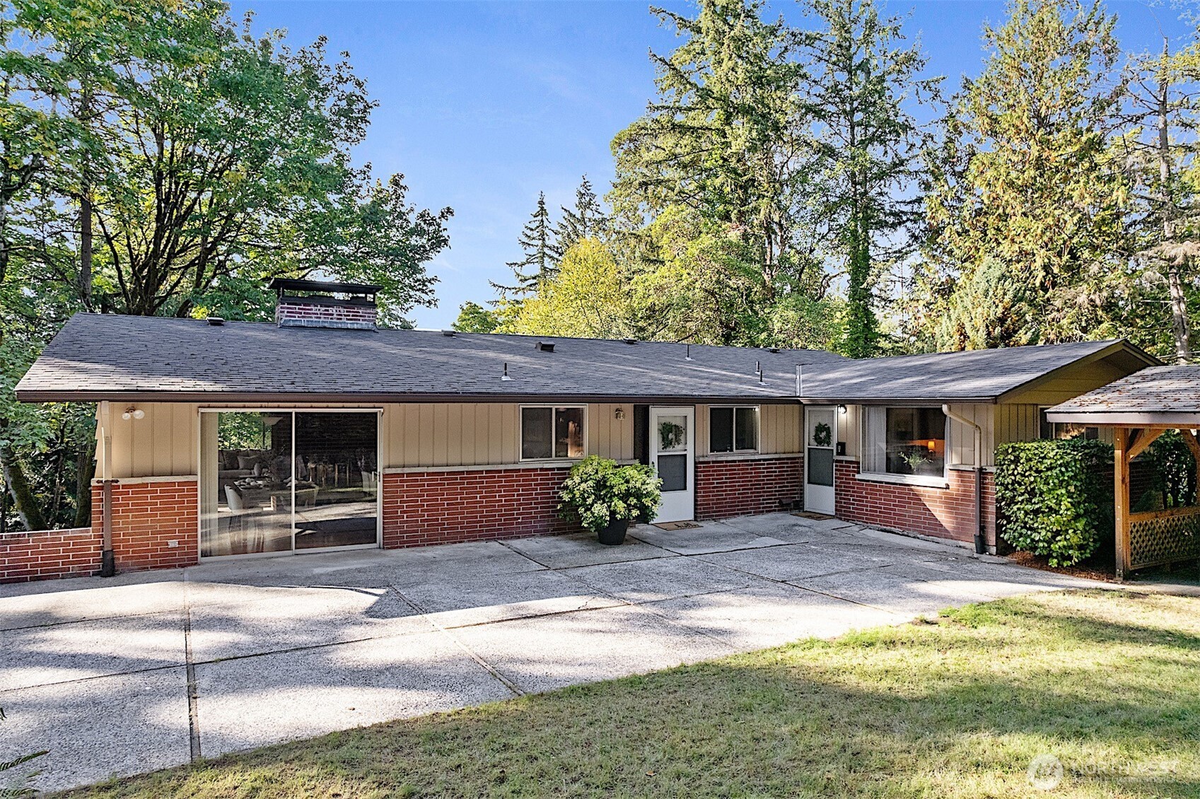 Single-story home with a red brick facade, broad concrete patio, and mature trees.