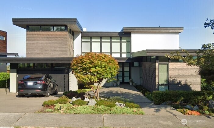 Modern two-story home with dark wood siding, broad windows, and colorful autumn landscaping.