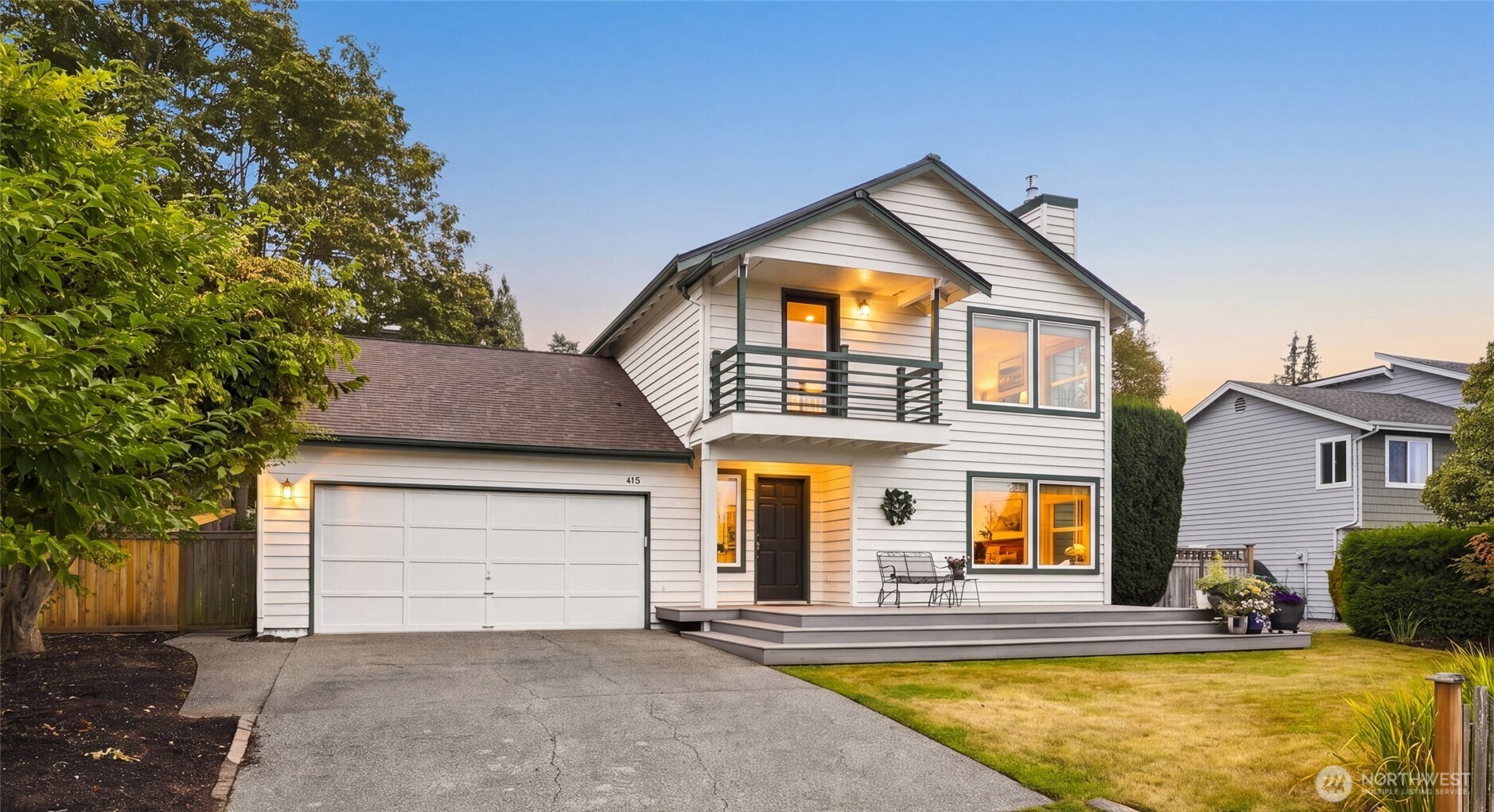 White two-story home with a green-railed balcony, attached garage, and warmly lit windows at sunset.