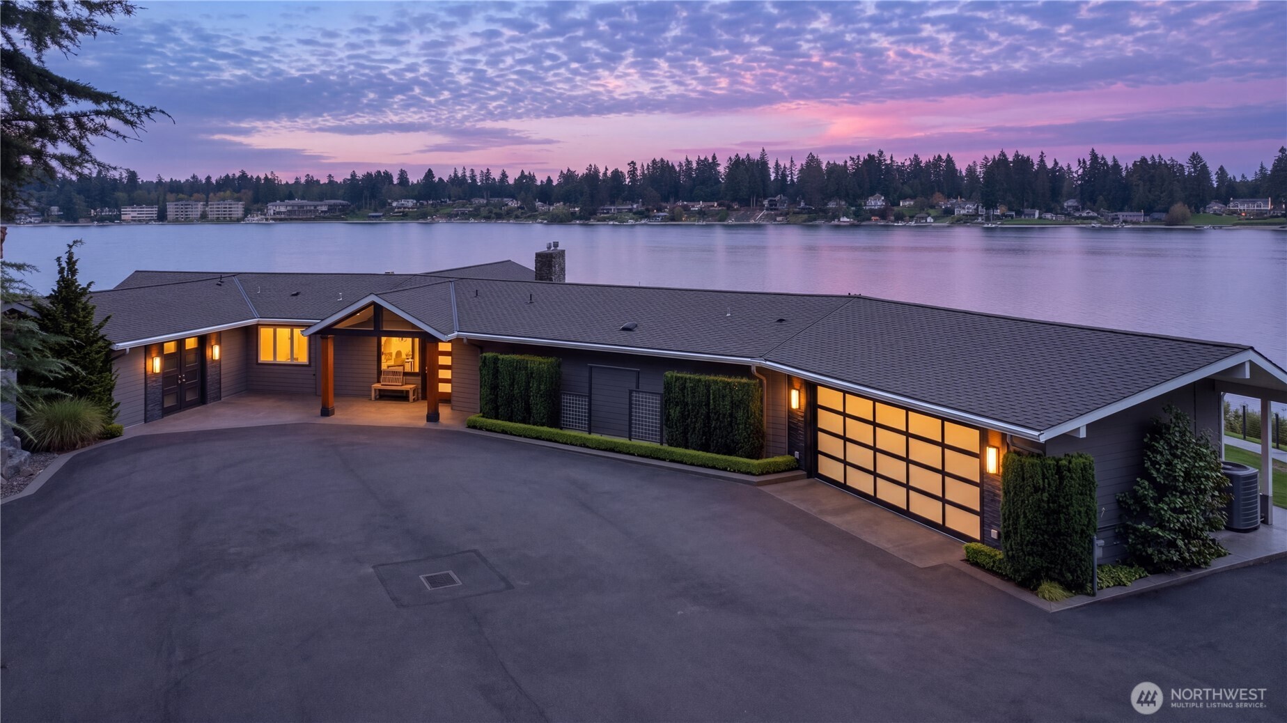 Modern lakefront home with an expansive driveway and warmly lit garage at dusk.