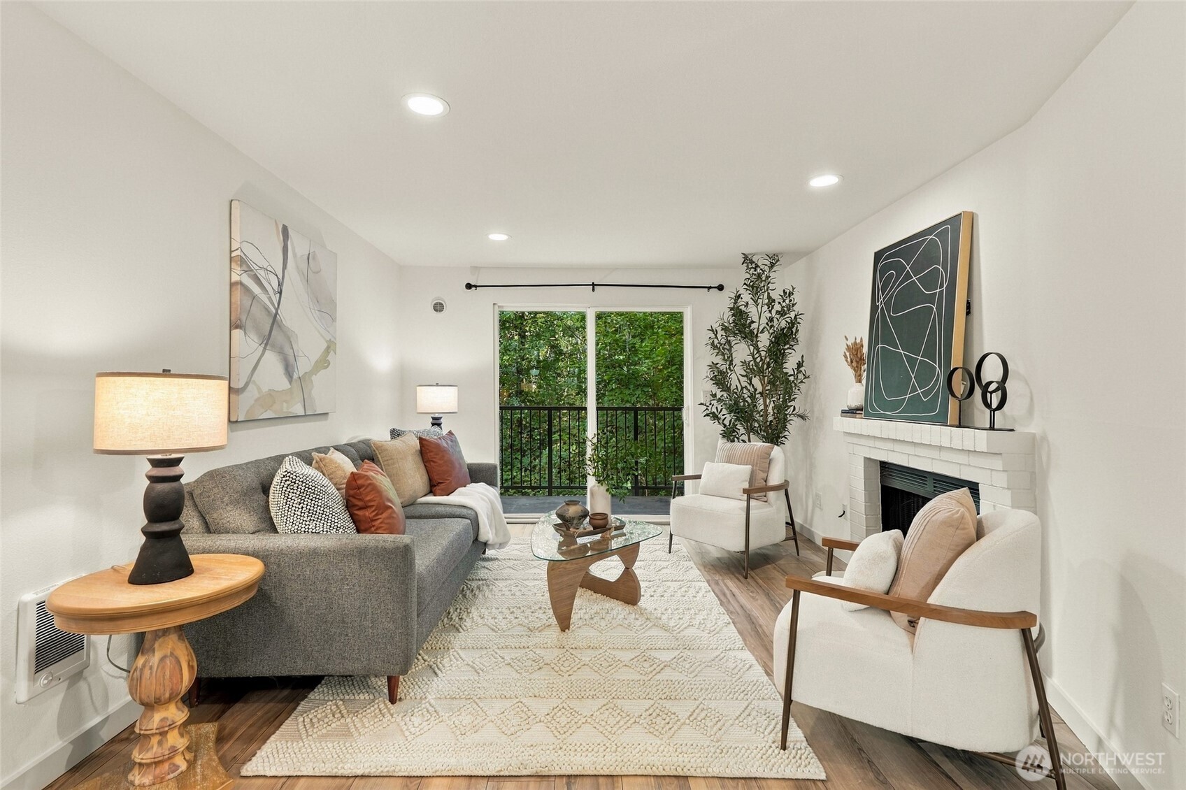 Staged living room with a gray sofa, cream armchairs, and a white brick fireplace beside leafy greenery.