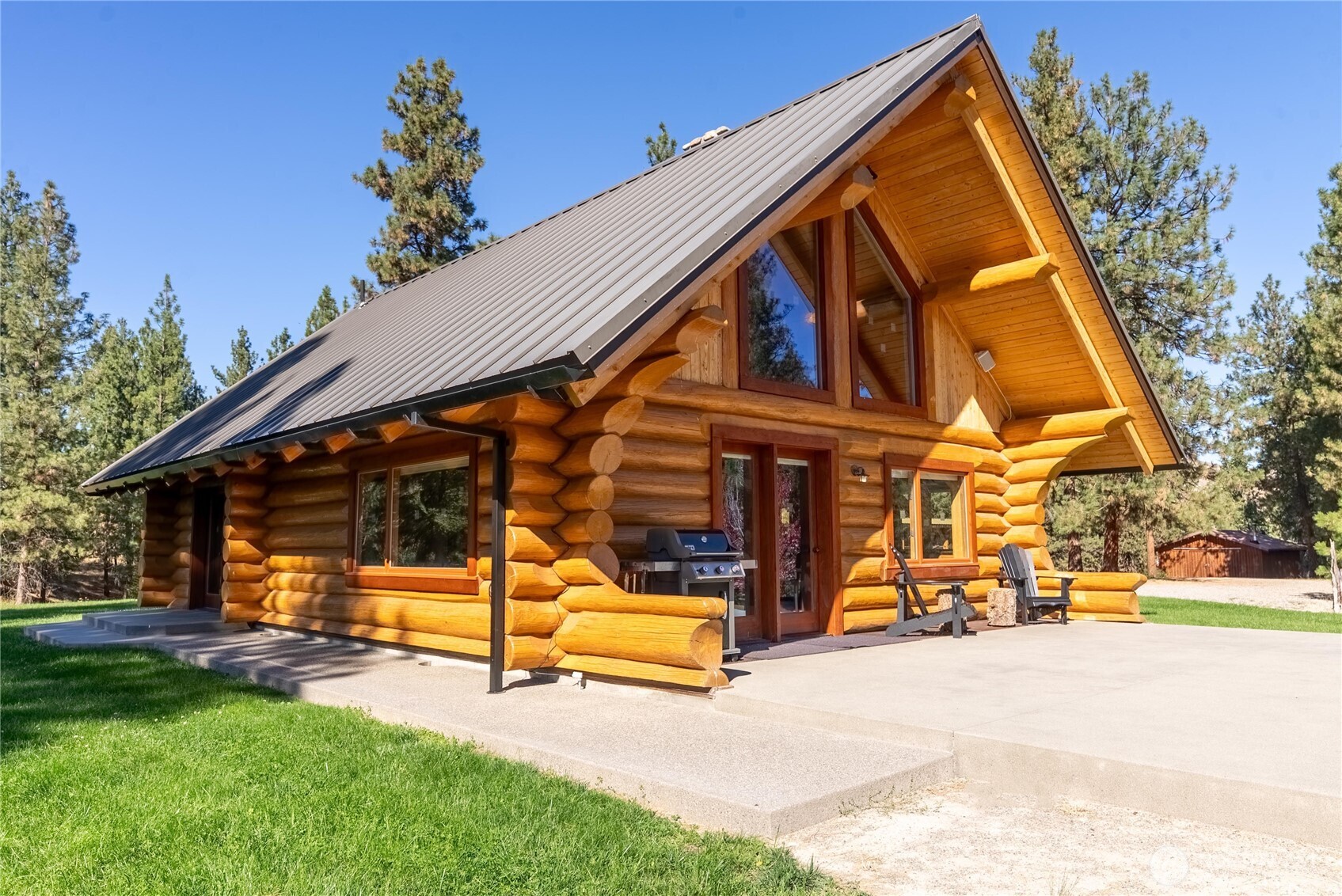 Rustic log cabin with a steep metal roof, concrete patio, and pine trees.