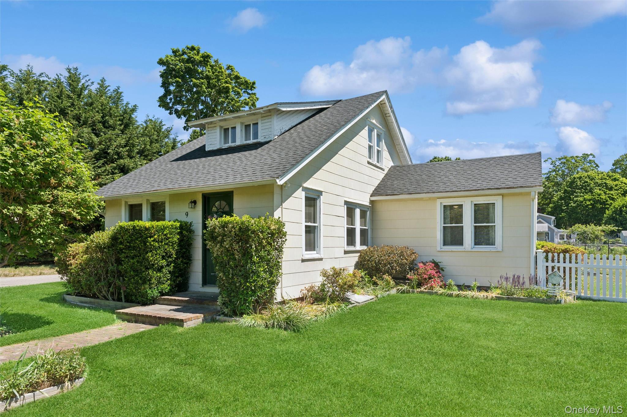 Light-colored house with a gabled roof, landscaped shrubs, broad green lawn, and white picket fence.