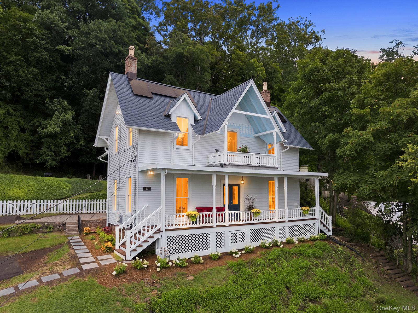 White two-story home with lit windows, wraparound porches, and a white picket fence among trees.