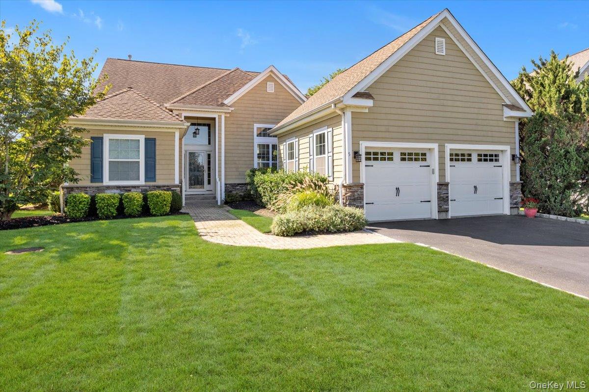 Tan shingle-style home with a two-car garage, landscaped shrubs, and a broad front lawn.