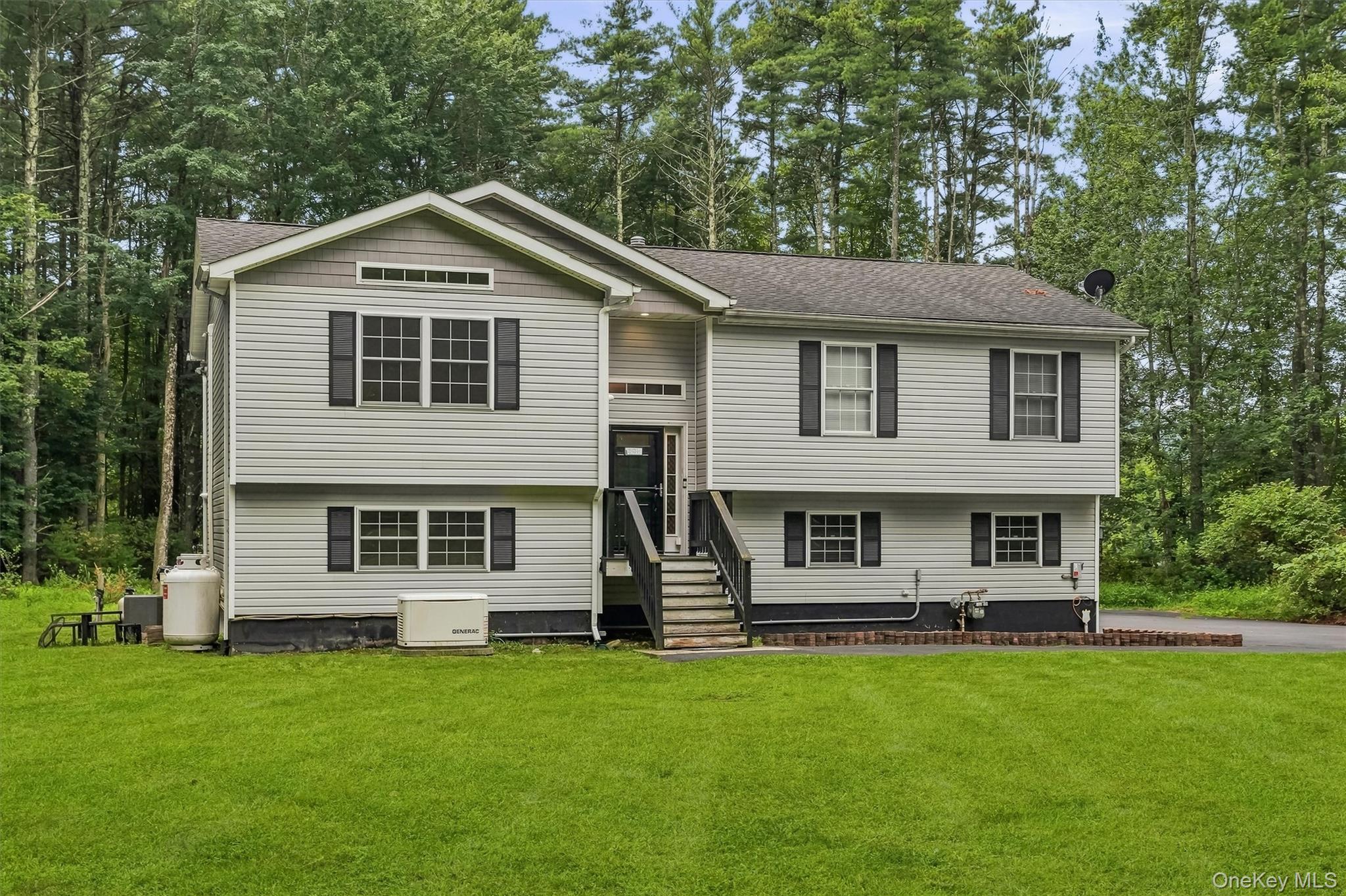 Gray split-level home with central stairs, a broad lawn, and dense woods behind it.