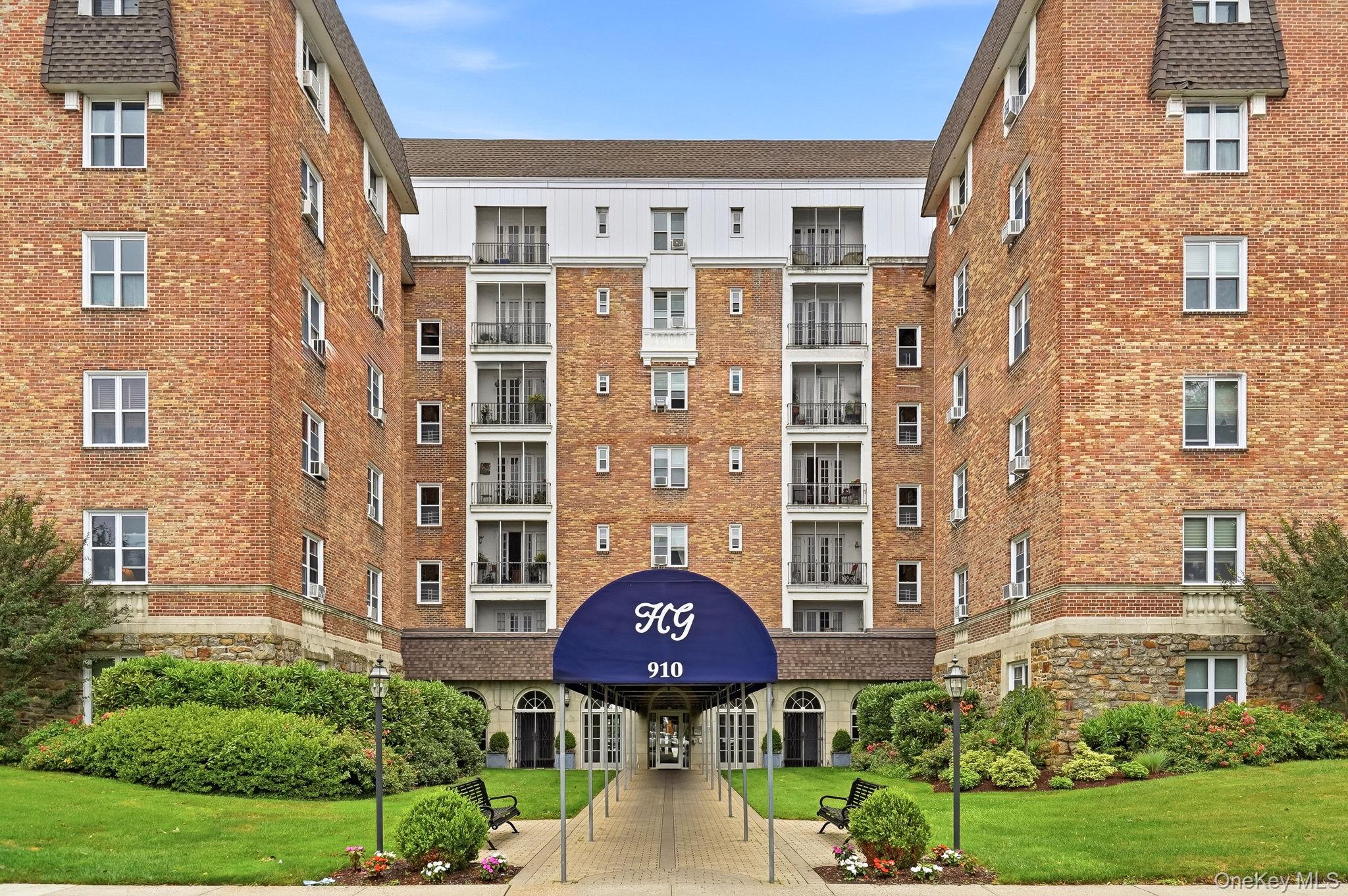 Brick apartment building with balconies, a blue entrance canopy, and landscaped grounds.