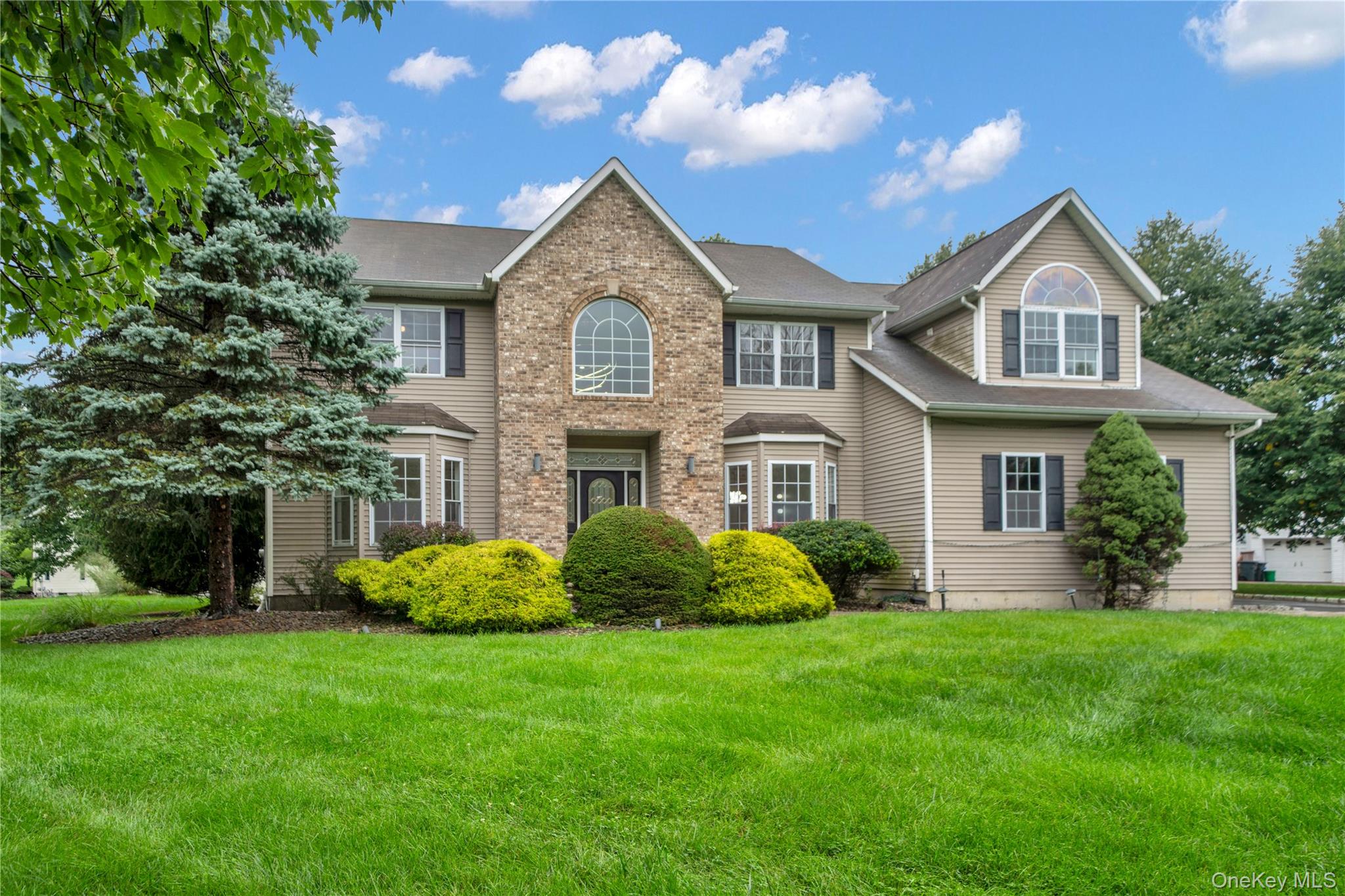 Two-story beige house with a brick facade, arched windows, landscaped shrubs, and a broad lawn.