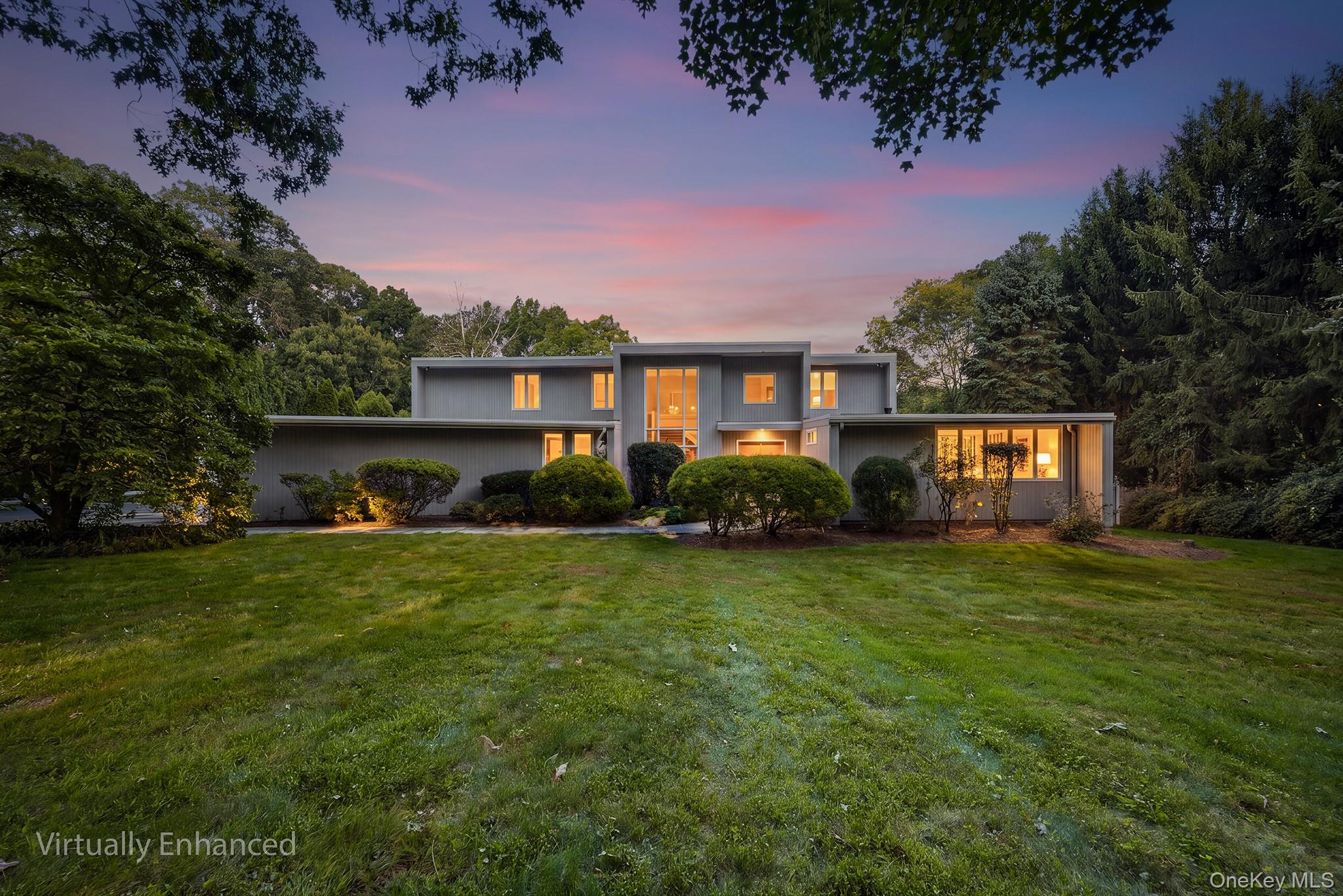 Modern gray home with illuminated windows, a broad lawn, and mature trees at dusk.