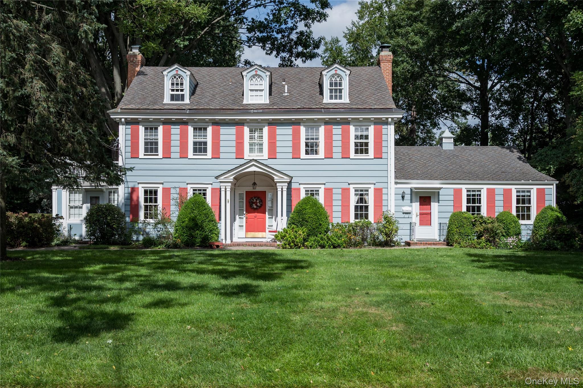 Light-blue two-story house with coral-red shutters, three dormers, and a broad green lawn.