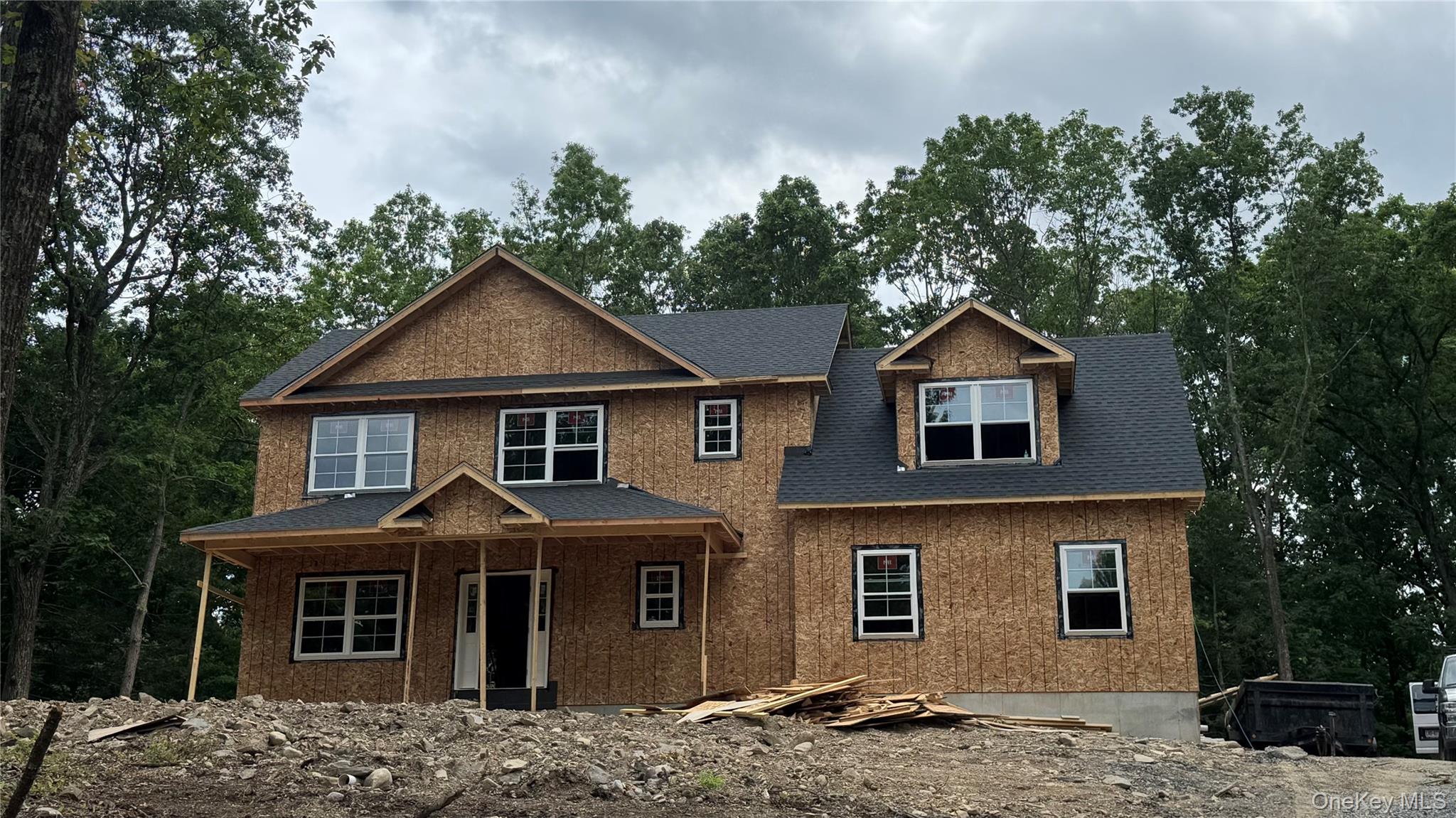Two-story house under construction with exposed sheathing, dark roof, and wooded surroundings.