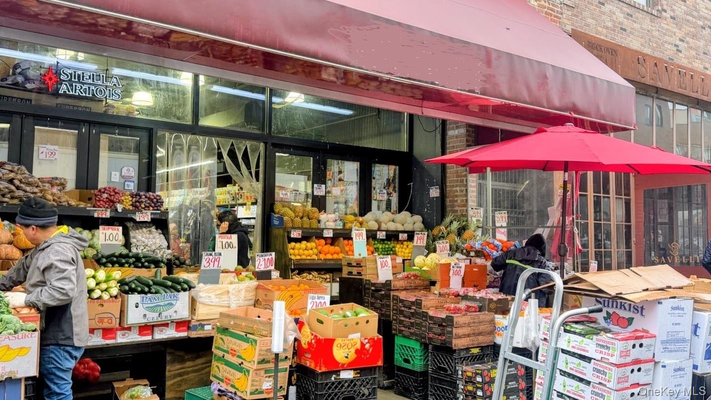 Sidewalk produce market with fruit and vegetables displayed beneath a red awning and umbrella.