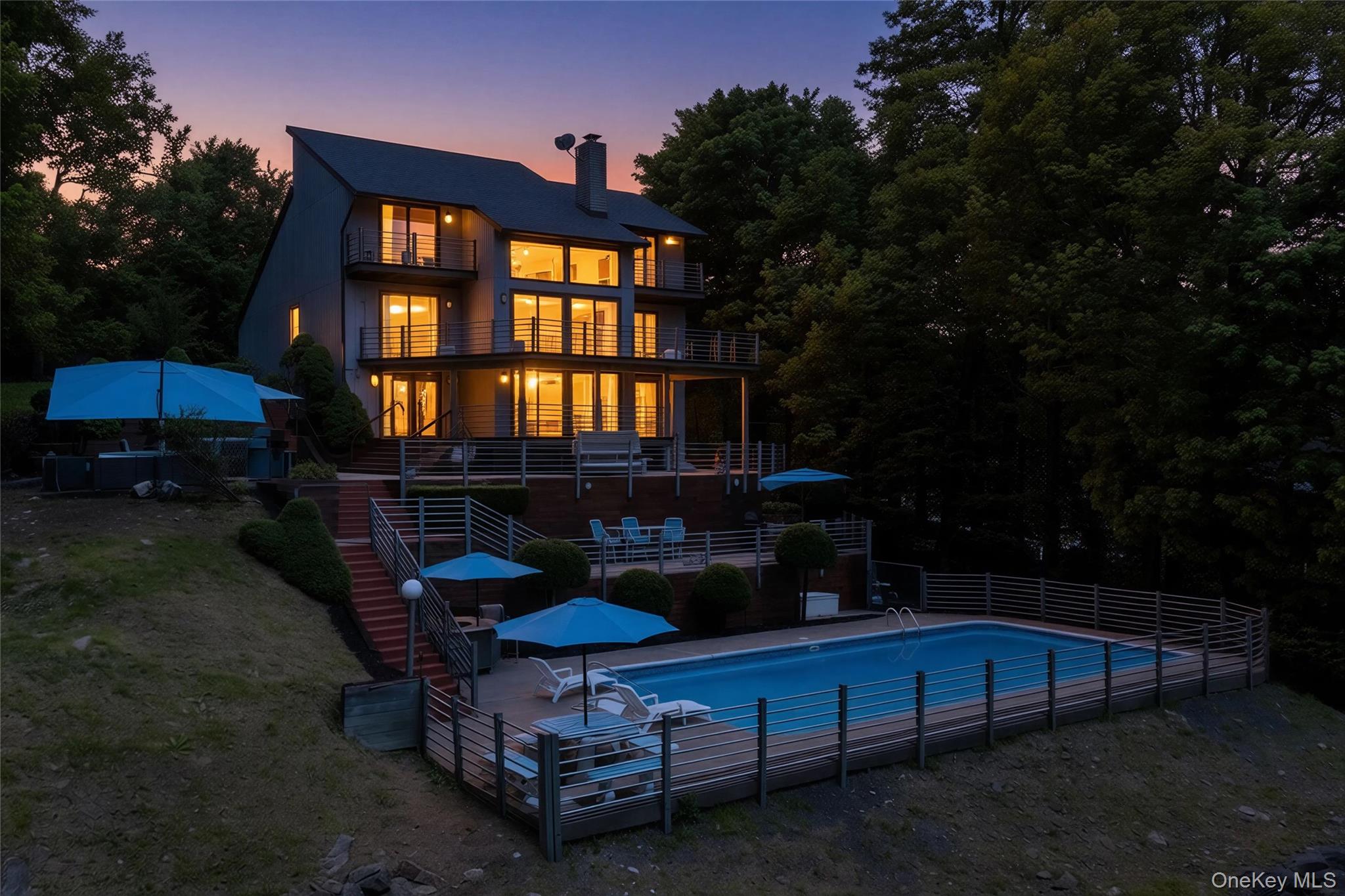 Modern multi-level home with illuminated windows and tiered decks overlooking a fenced swimming pool.