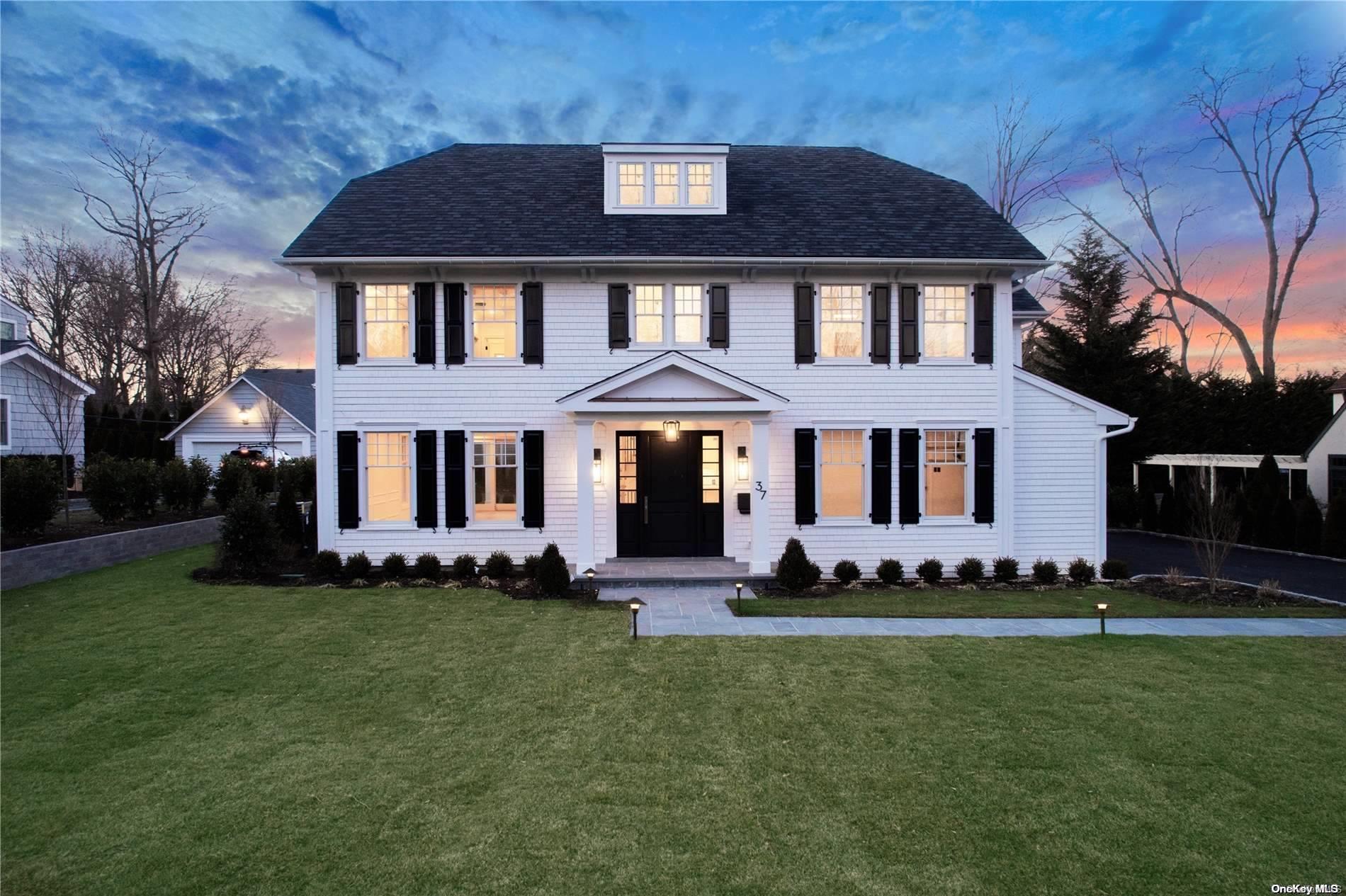 White two-story house with black shutters and glowing windows behind a broad lawn at dusk.