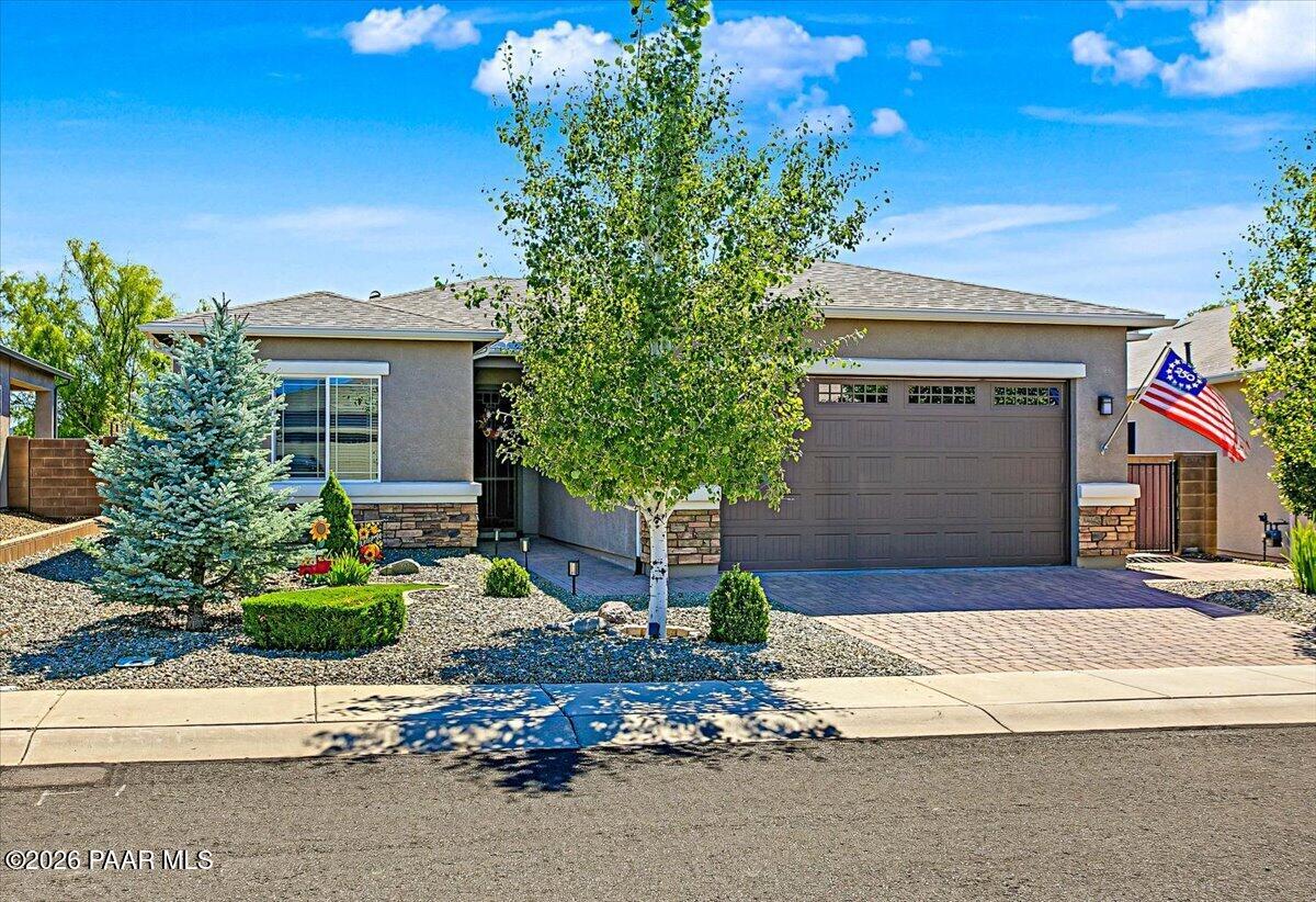 Single-story gray stucco home with a two-car garage, gravel landscaping, and a large leafy front-yard tree.