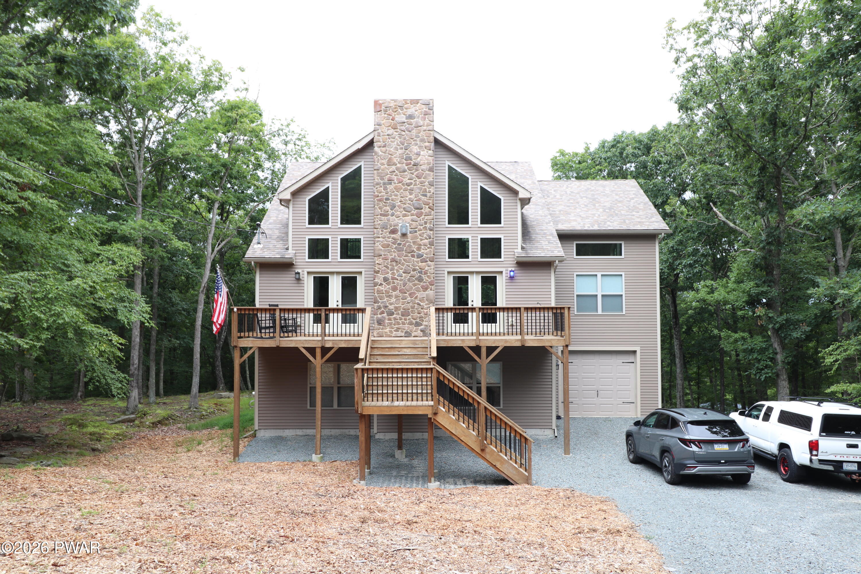 Beige-sided house with a tall stone chimney, raised deck, and gravel driveway among mature trees.