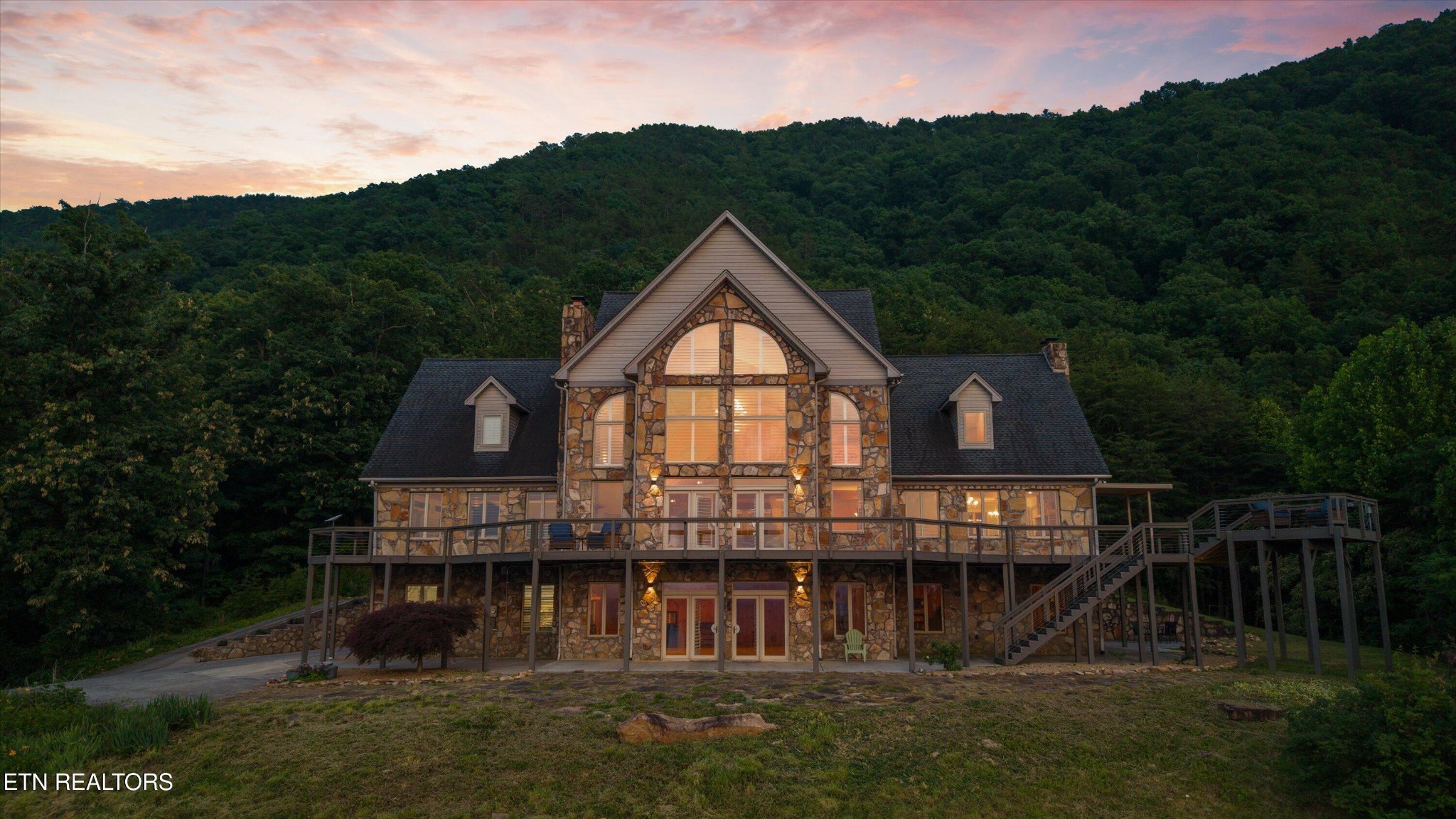 Stone house with broad decks and glowing windows beneath a wooded mountain ridge at dusk.