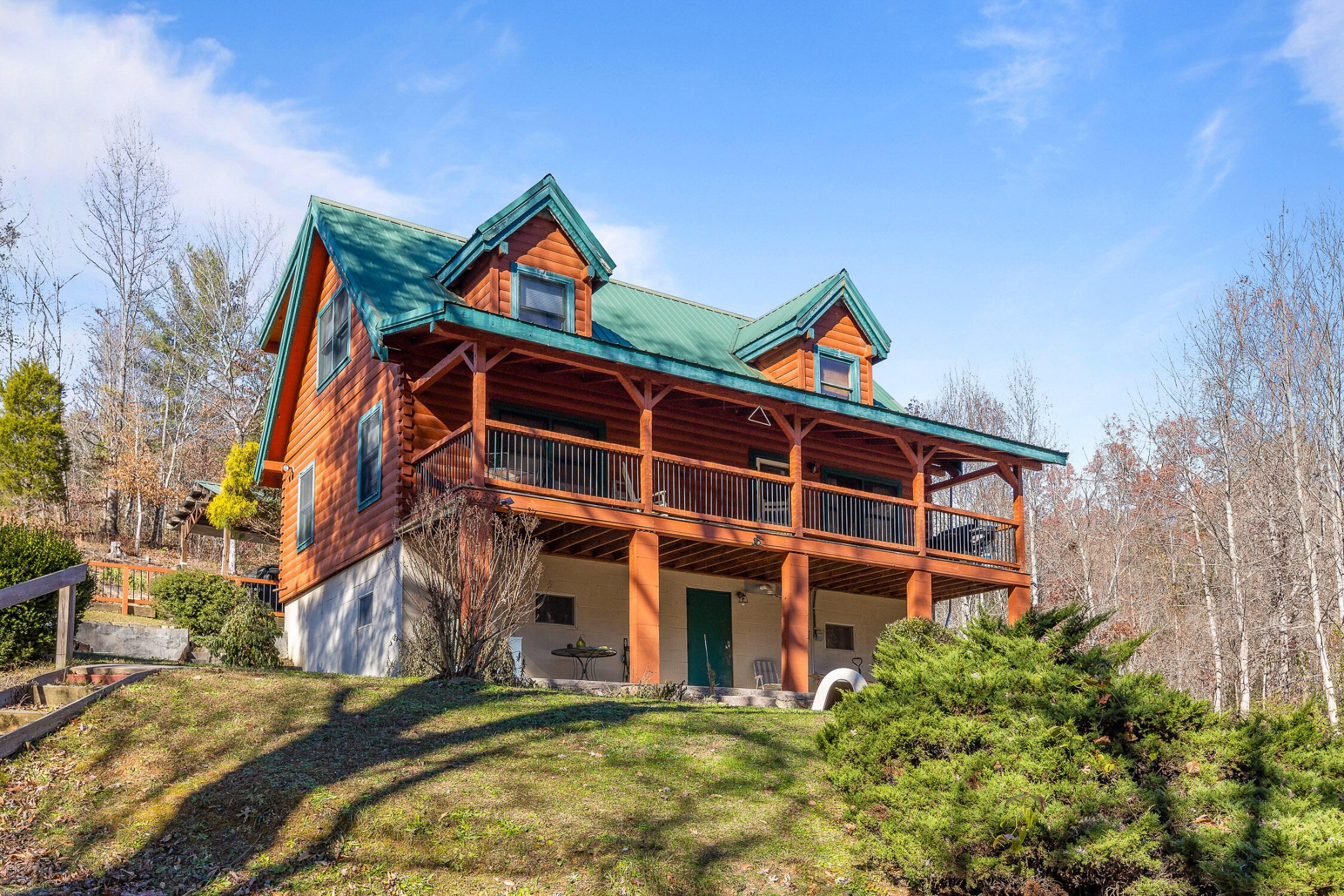 Log cabin with a green metal roof and long covered porch on a wooded slope.